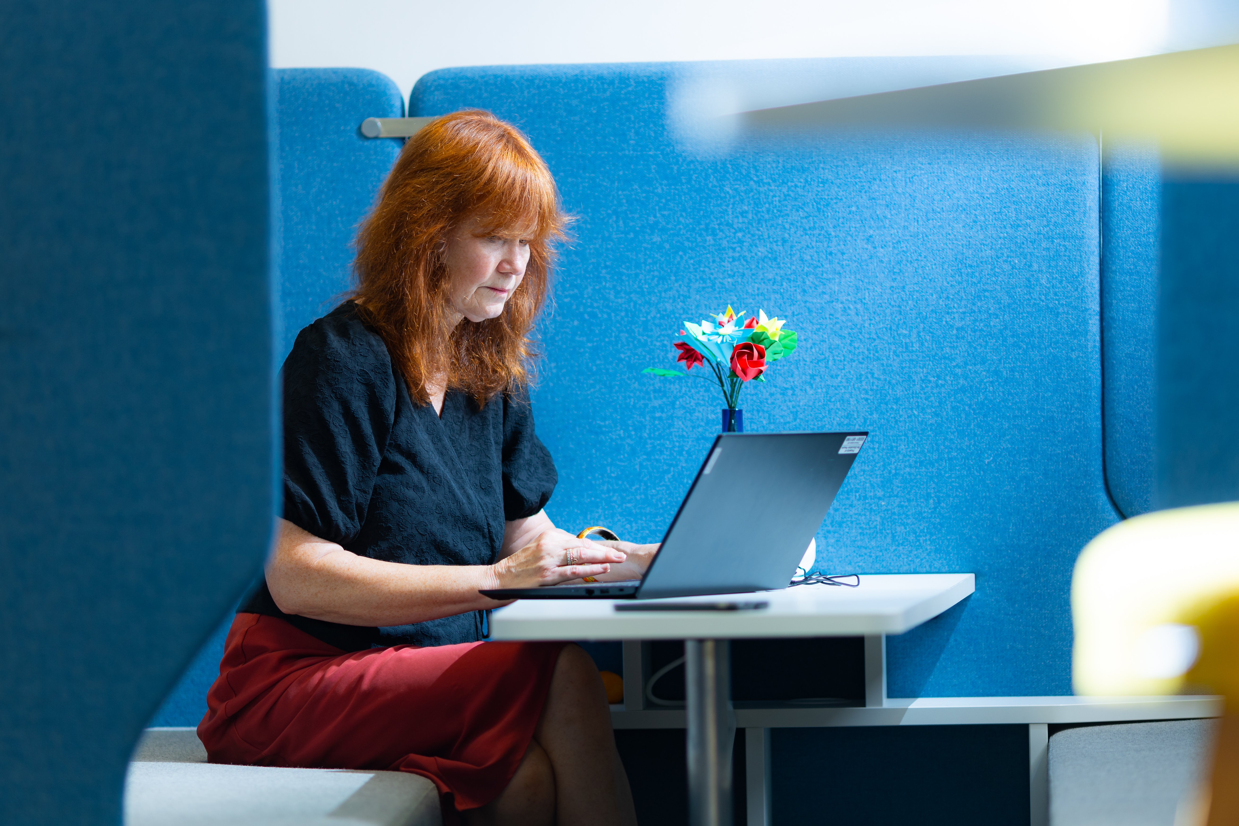 ALT - A woman with red hair sits at a blue booth, focused on her laptop.
