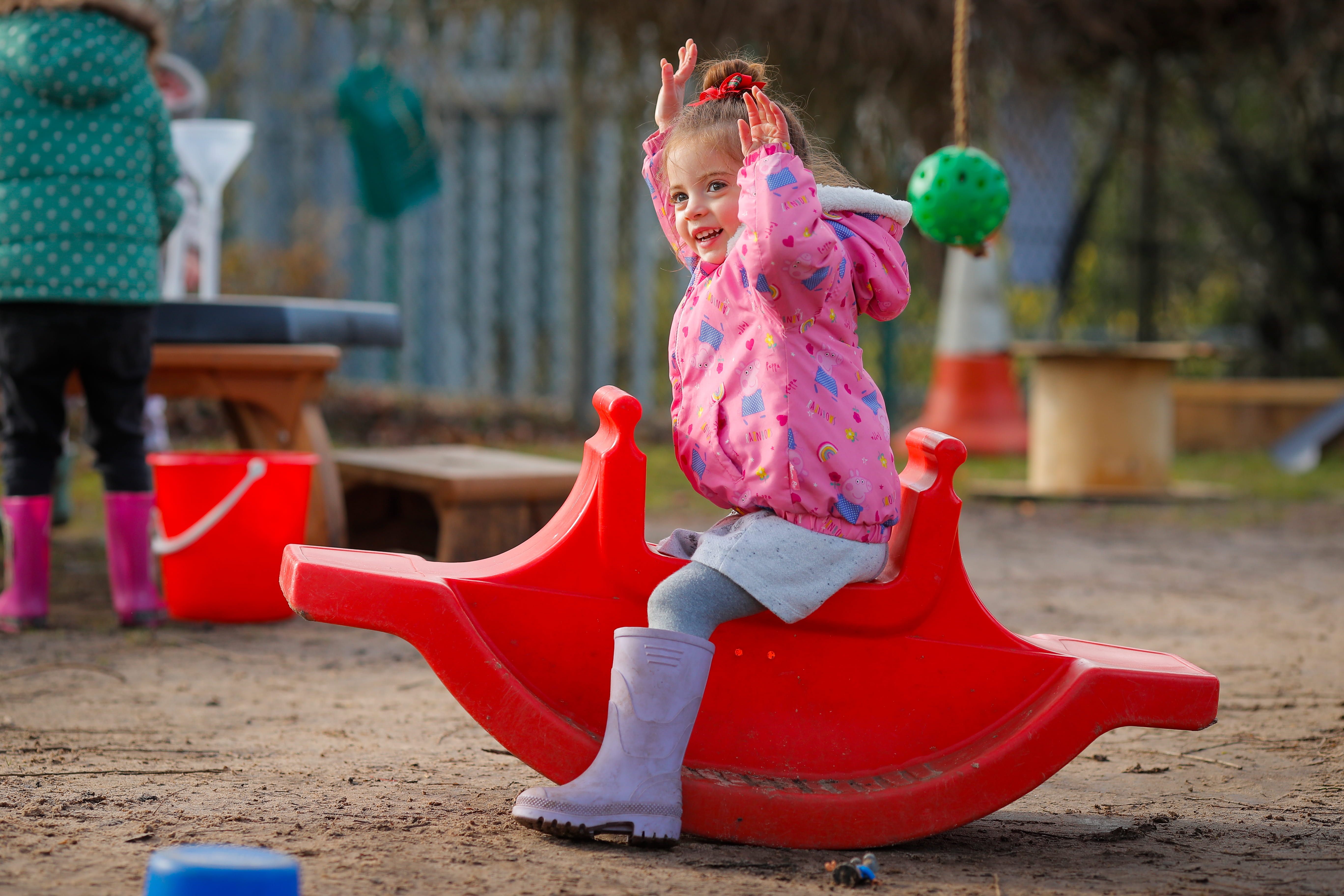 ALT - A smiling young girl in a pink jacket and purple boots plays on a red rocking toy in a playground.