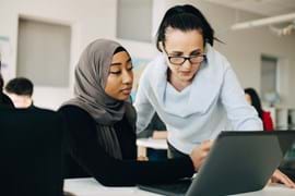 ALT - Lady sitting at a laptop, other lady standing looking at the screen having a conversation