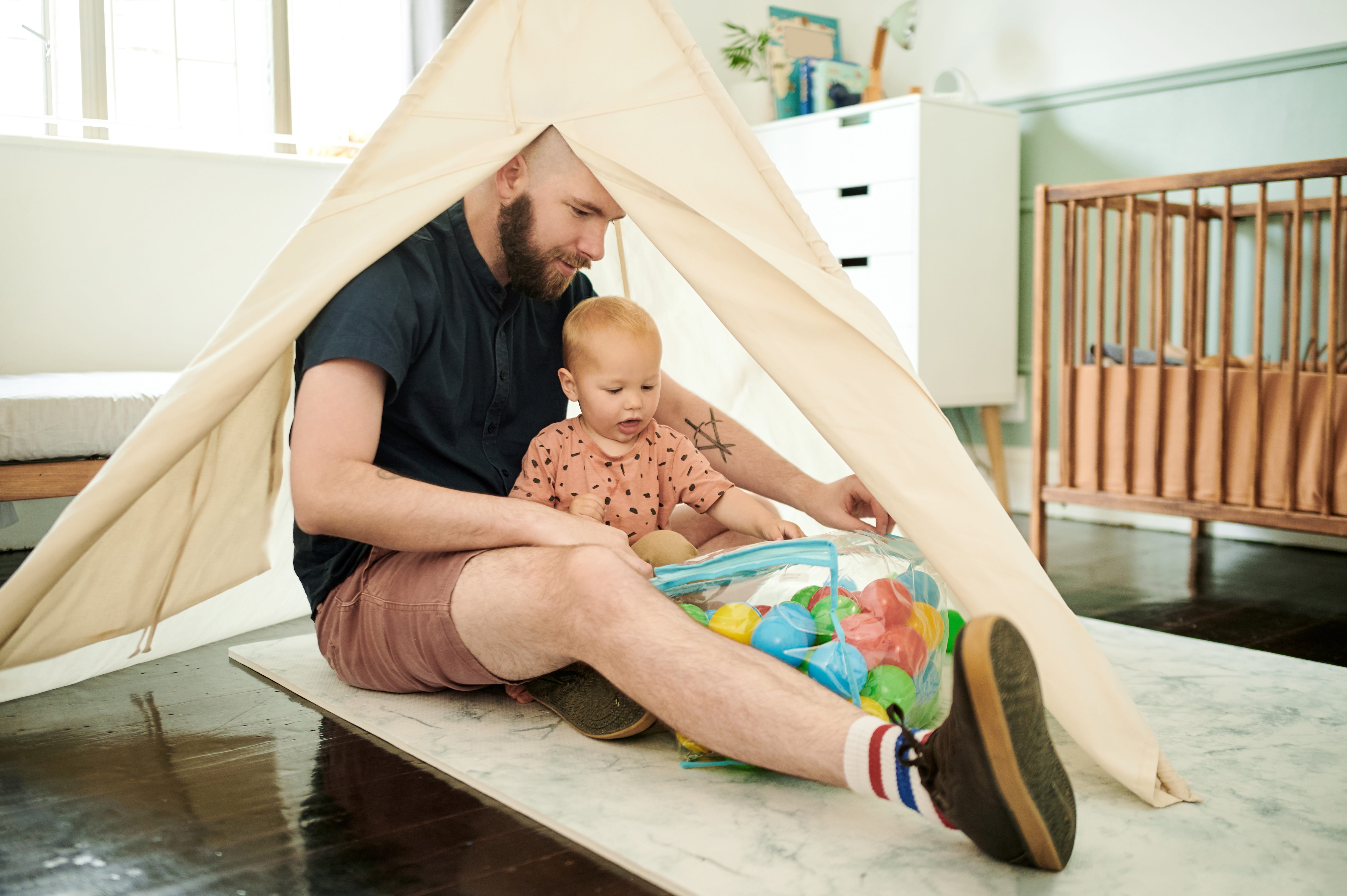 A father and toddler sit together under a small play tent, surrounded by colourful toys, in a cosy, softly lit room
