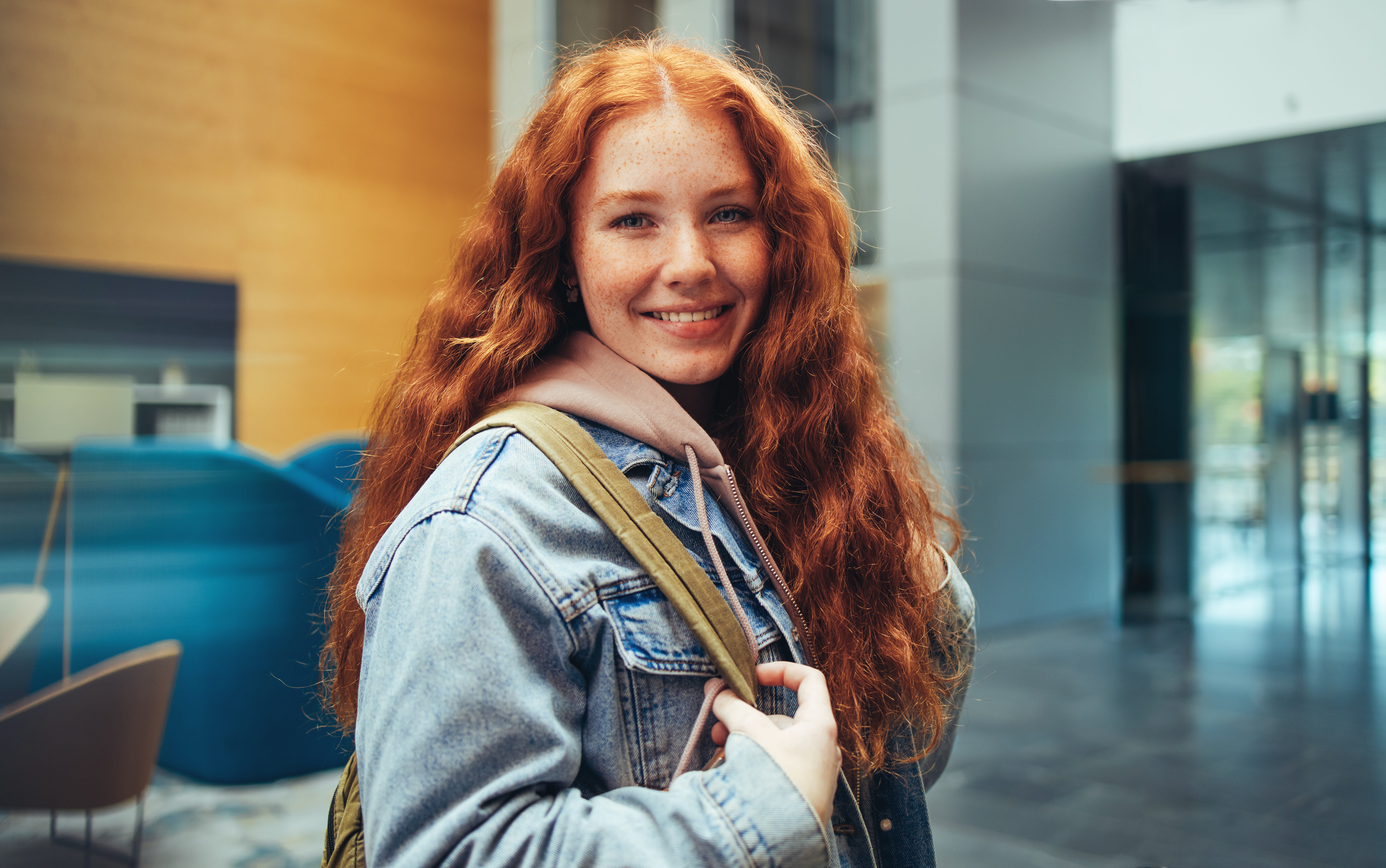 ALT - Smiling young woman with long red hair and freckles, wearing a denim jacket and backpack.