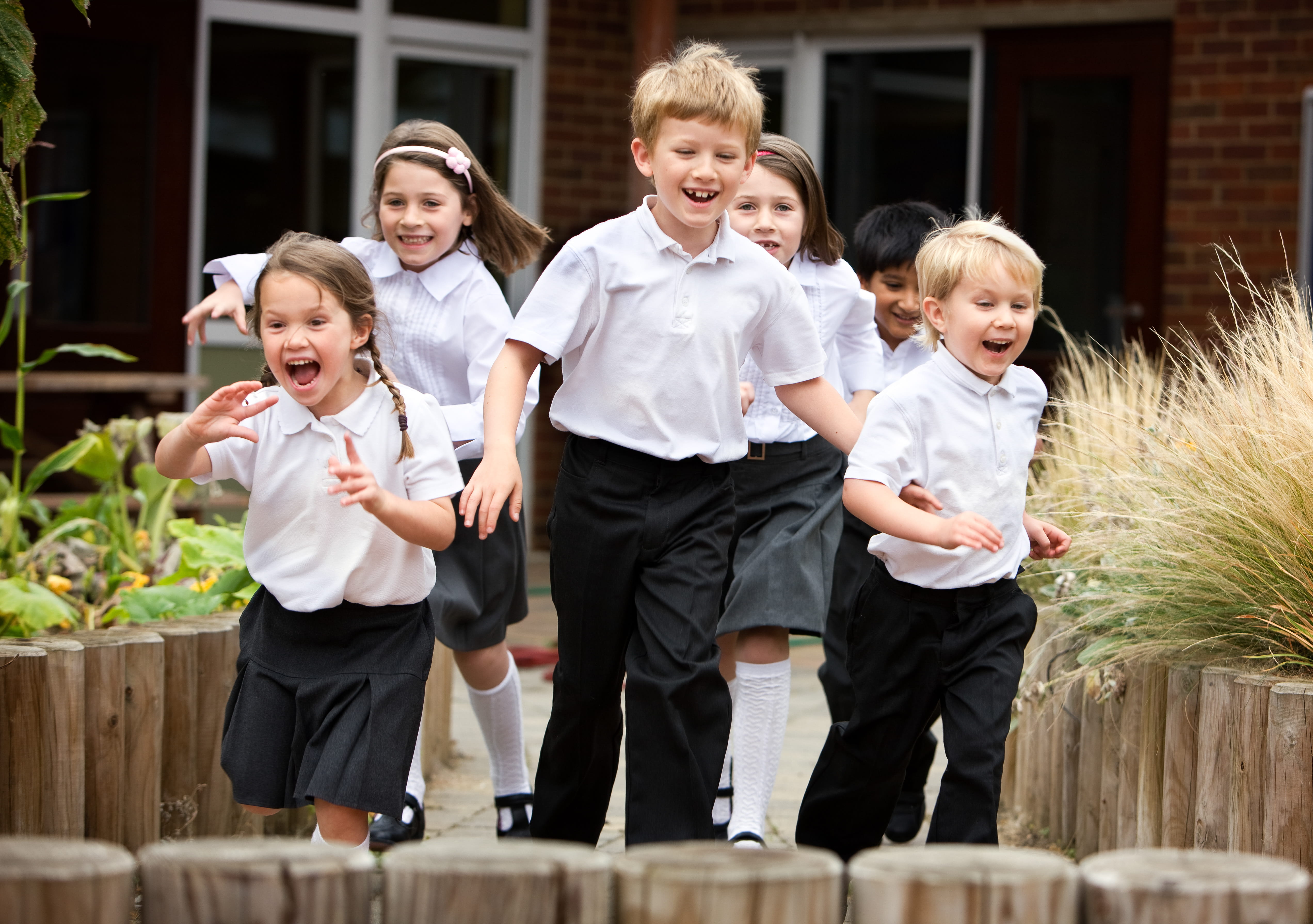 Six children in uniform joyfully run on a schoolyard path, surrounded by greenery.