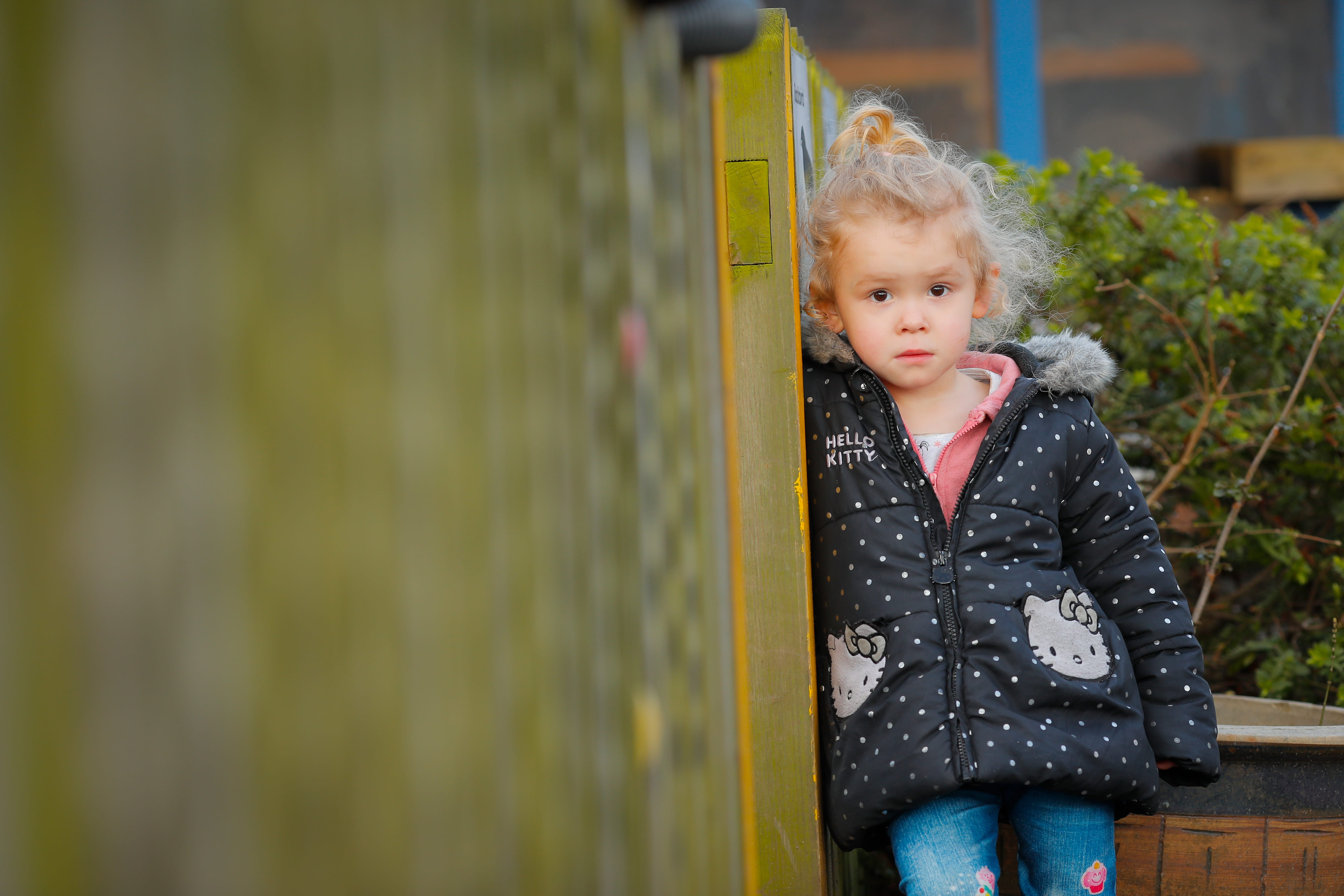 A young child with curly hair, wearing a black Hello Kitty jacket, leans against a wall outdoors.
