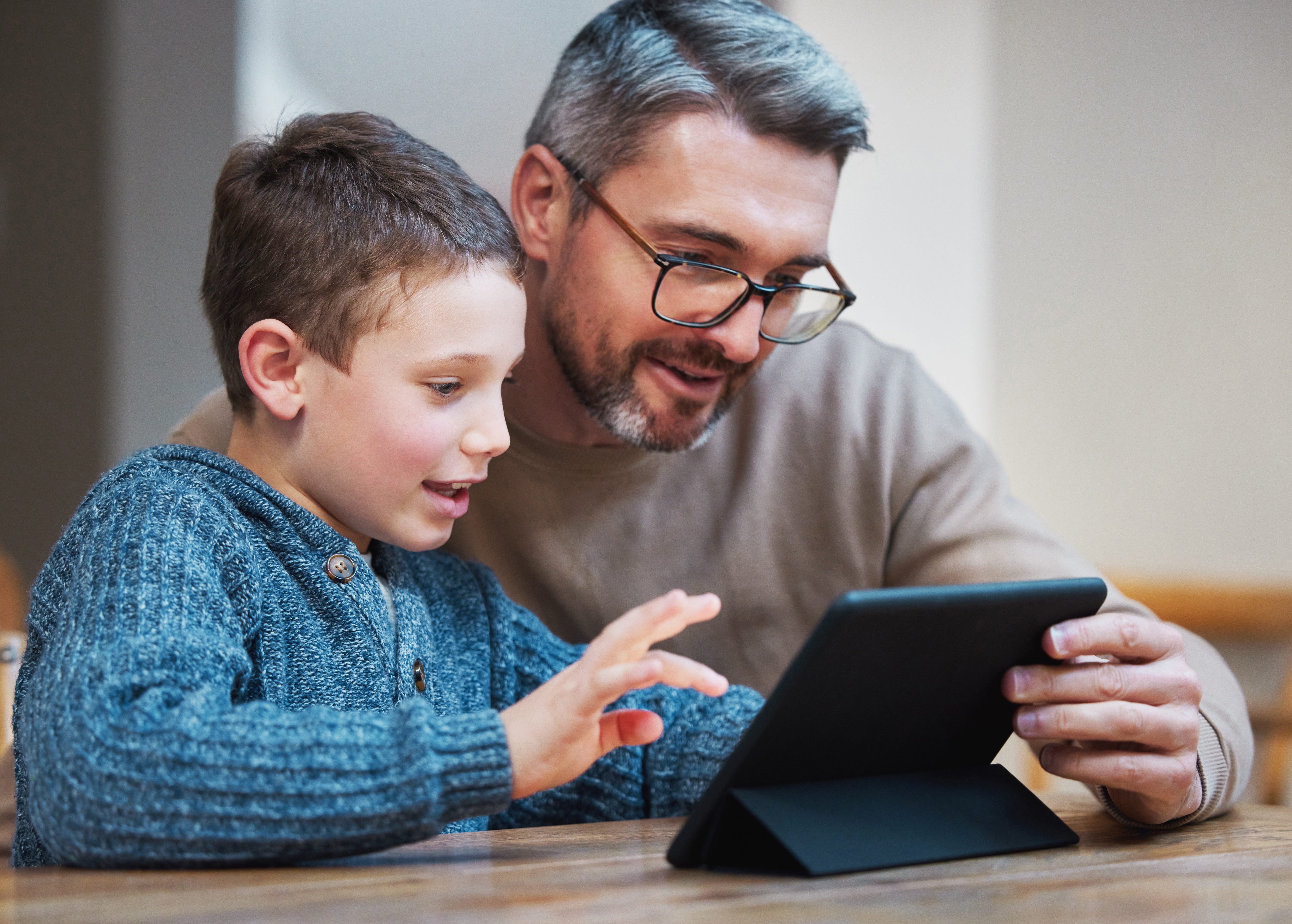 ALT - a dad and child sitting at a table dad is holding an ipad child is looking at screen and using his finger to touch the screen