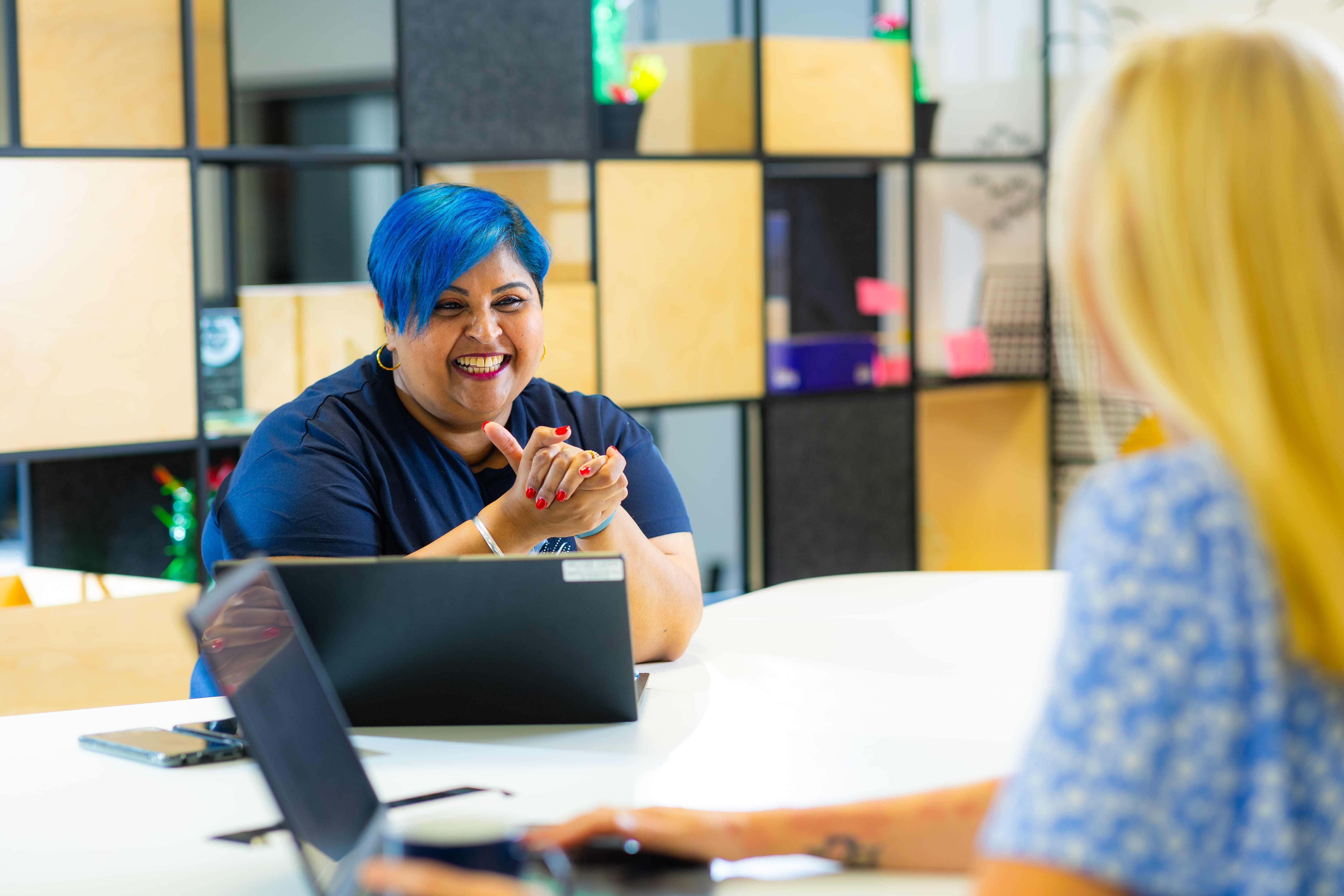 ALT - two ladies sitting at a desk working on laptops having a conversation