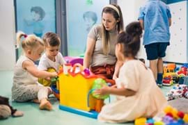 ALT - an adult sitting on the floor with nursery children watching them whilst they play with toys