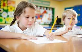 Two children sitting at a desk one child is writing with a pencil, the other is thinking resting his pencil on his head