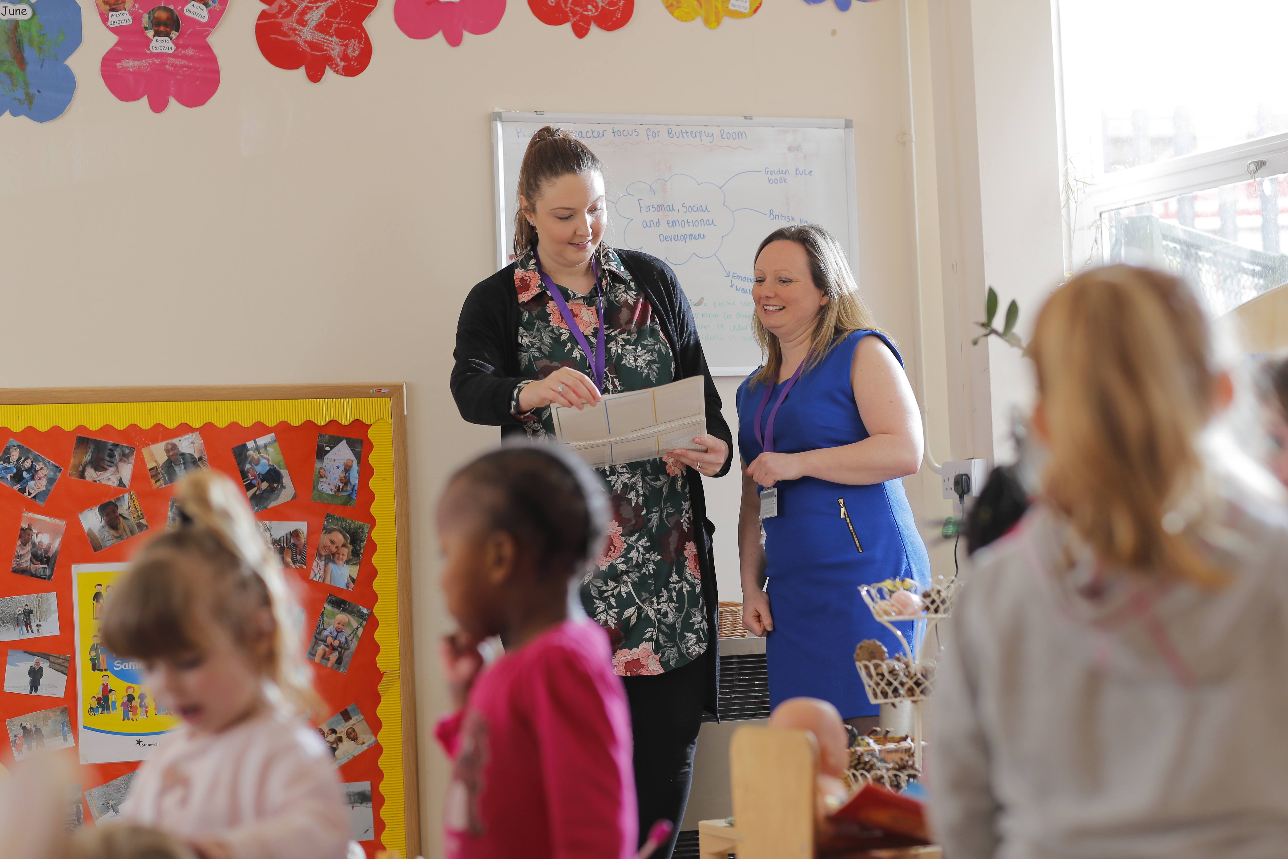 Two ladies talking and smiling looking at a piece of paper next to a board with photos on, there are children in the foreground playing