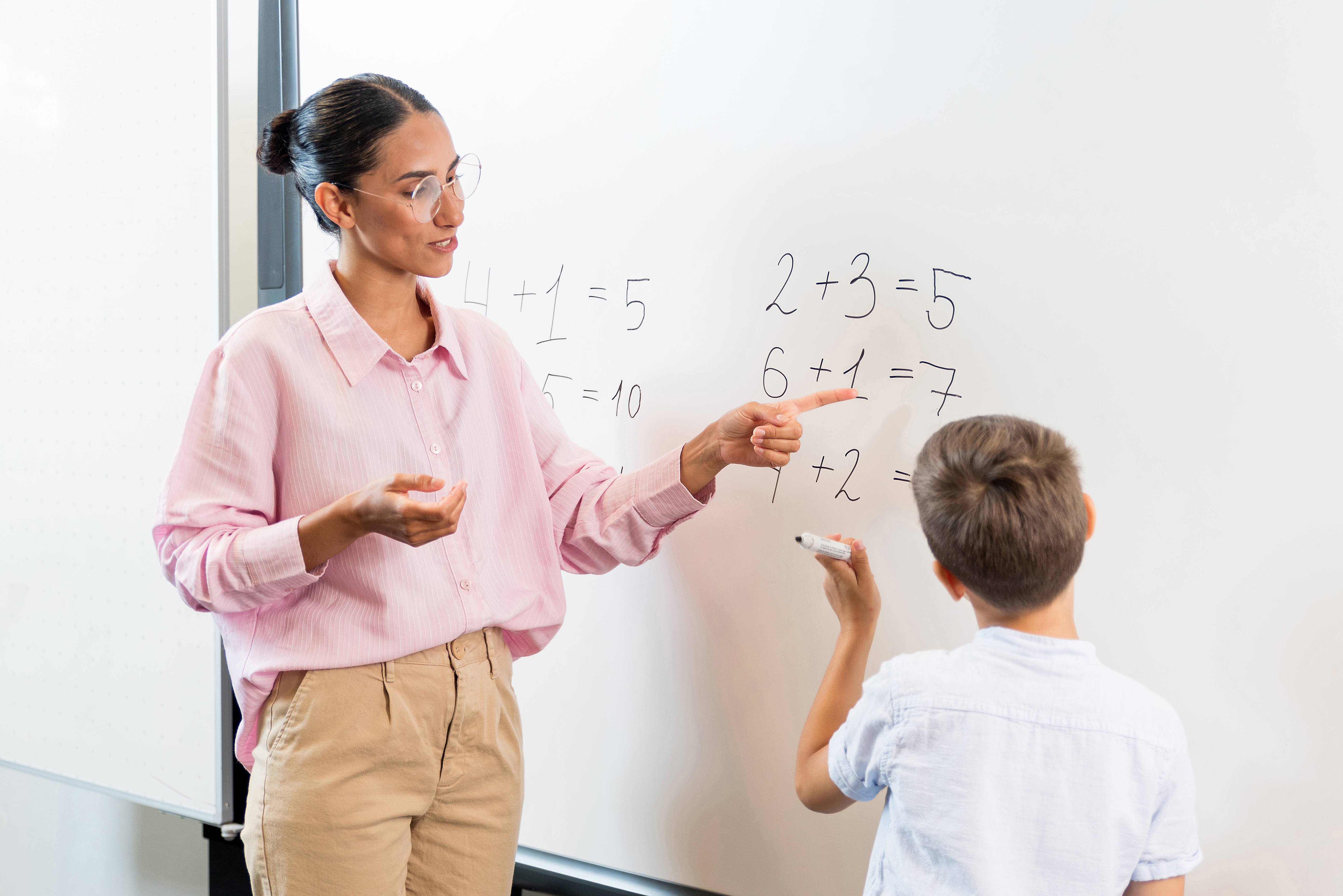 Teacher and child standing next to white board, child has a pen in his hand whilst the teacher points at numbers on the board