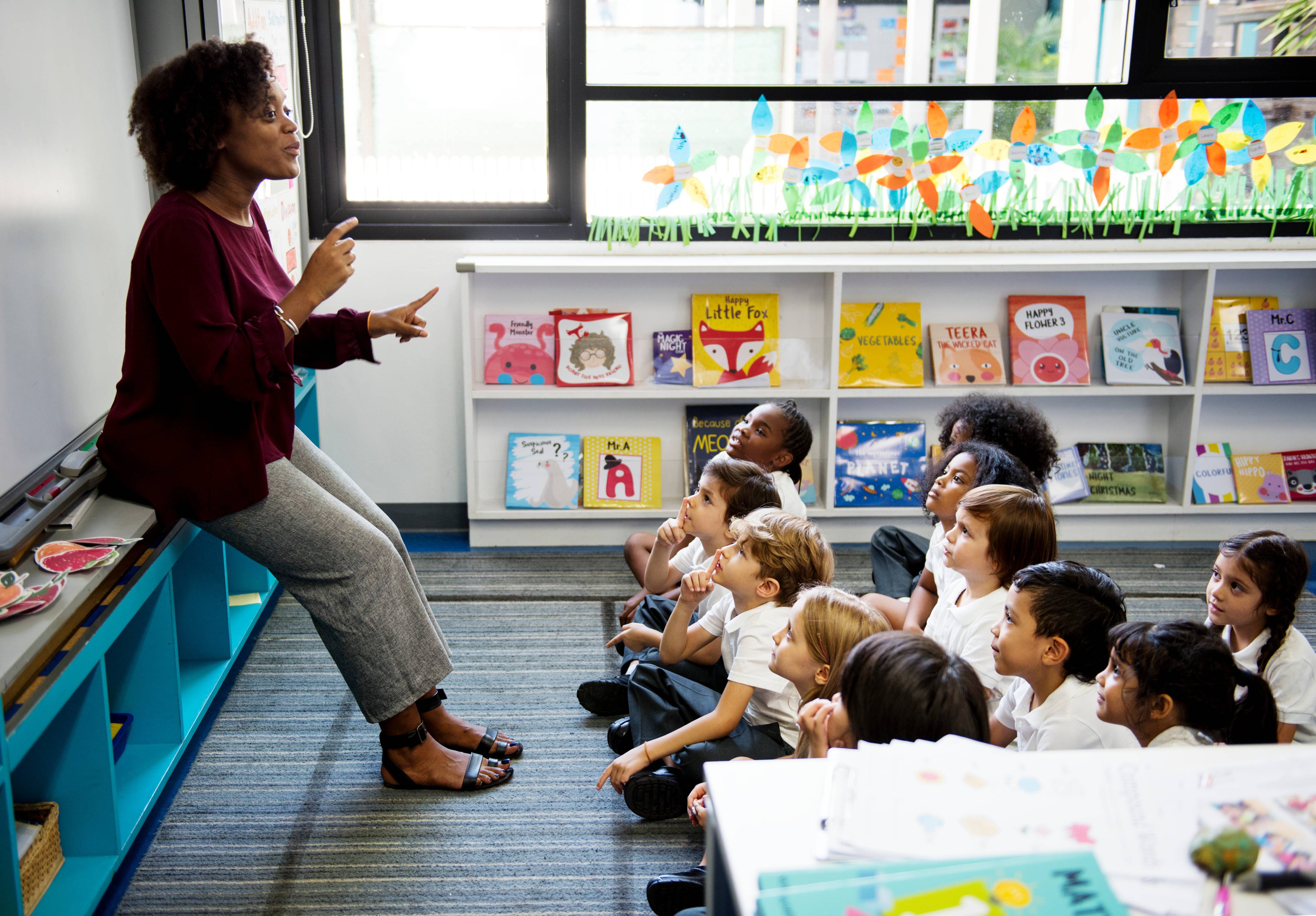 ALT - teacher standing at the front of the class, children sitting on the floor looking up at her with their finger on their lips