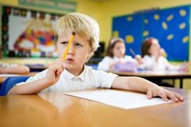 Child sitting at a desk with pencil in hand pointing it at his head and thinking, other children are blurred in the background