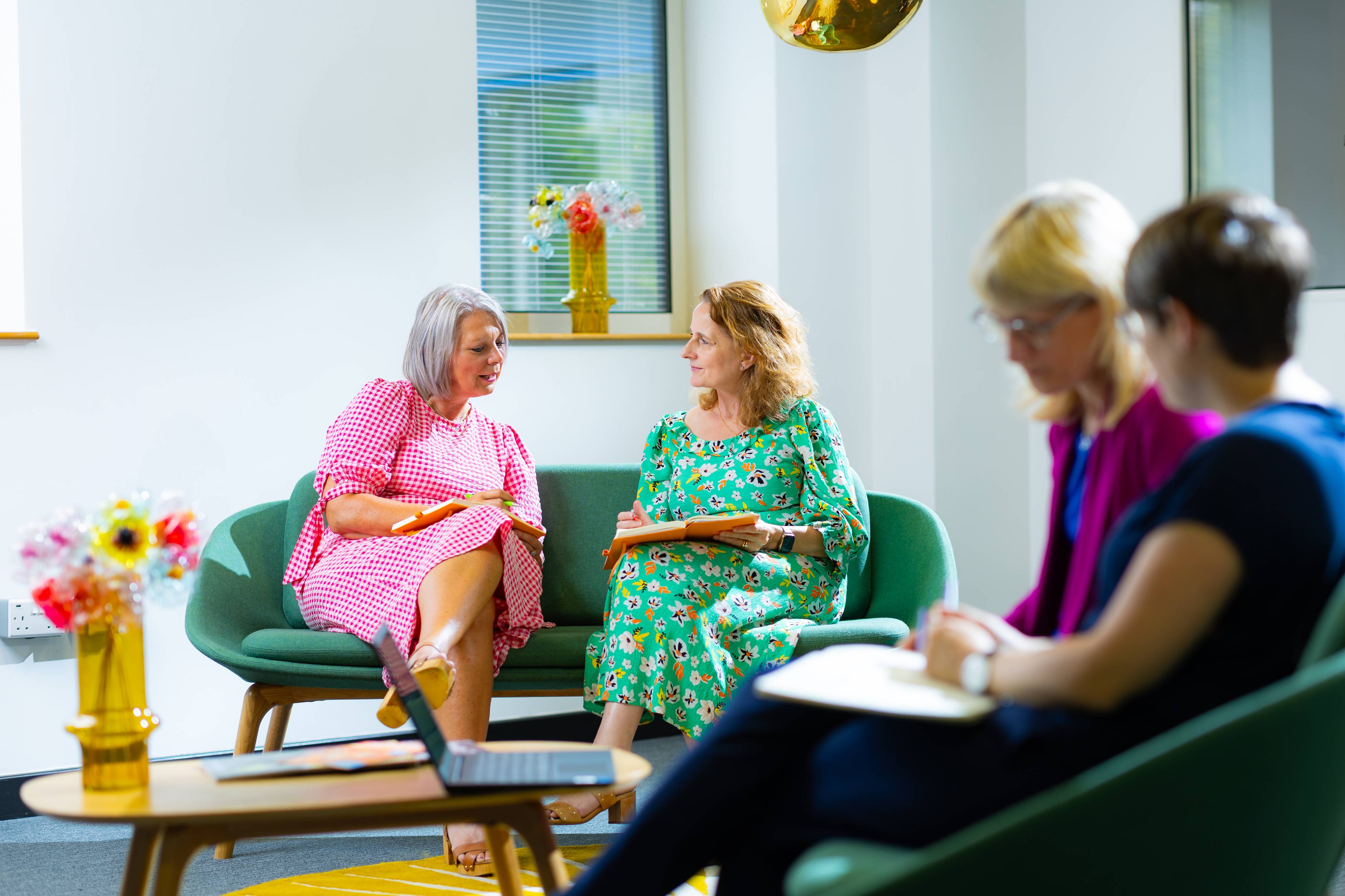 Two ladies sitting on a sofa holding notepads and talking to each other there are two other ladies sitting on sofas in the foreground one has a notepad. There is a laptop on the table in front of them with a vase of flowers.
