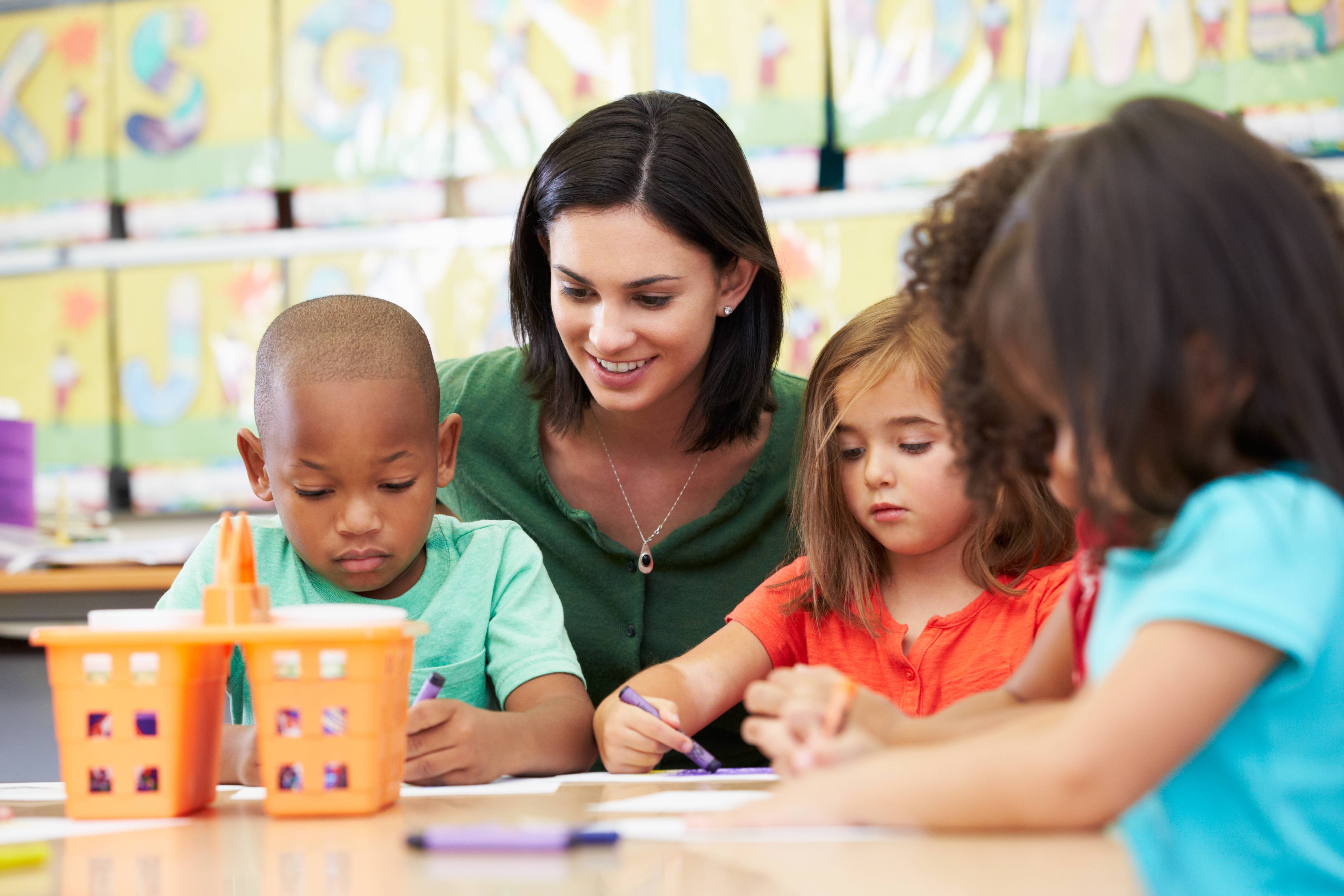 Teacher squatting behind children sitting at a table, looking over their shoulder watching them colour and draw with crayons.