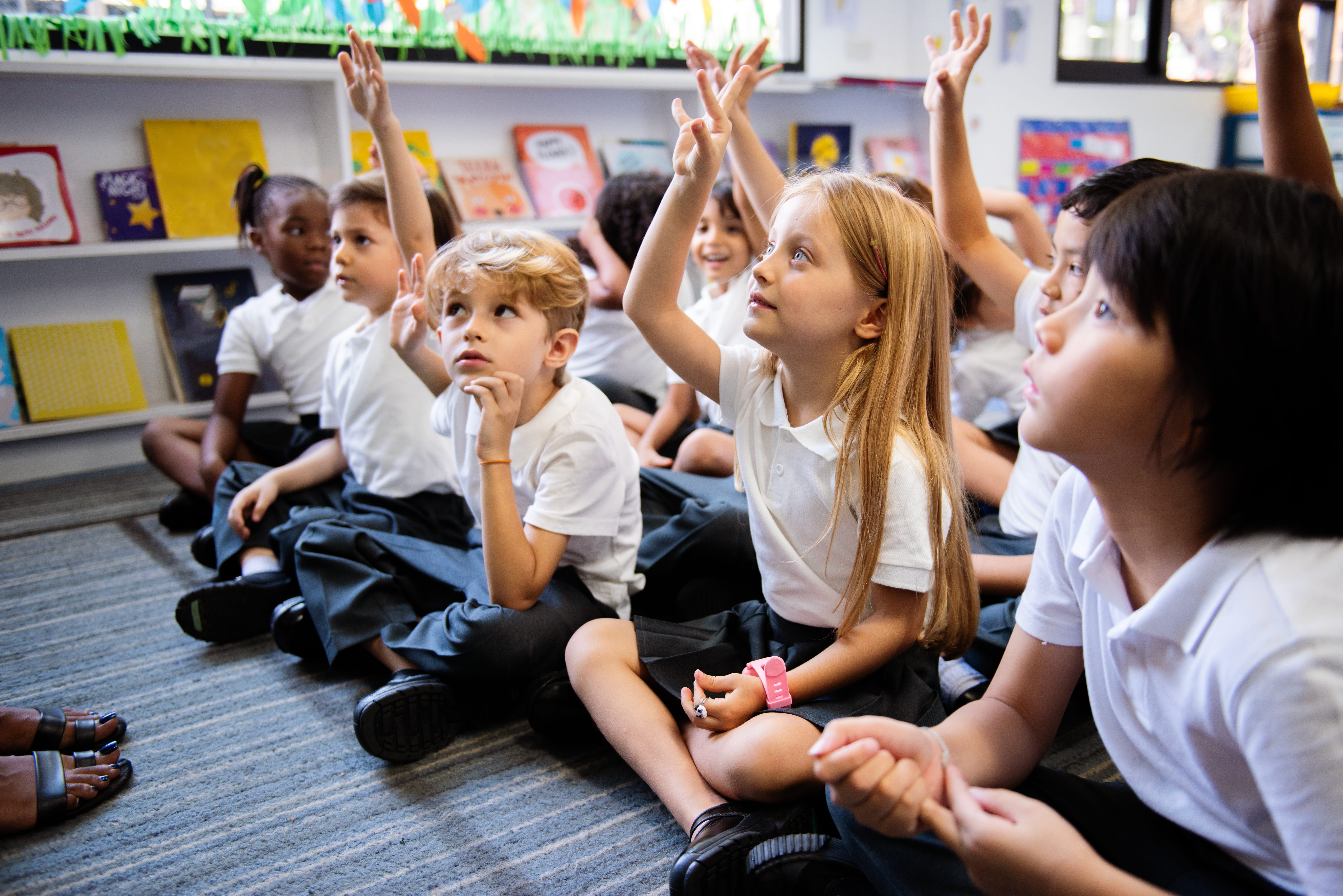 Children sitting on the floor in a classroom, holding their hands in the air to answer questions there are books on shelves in the background.