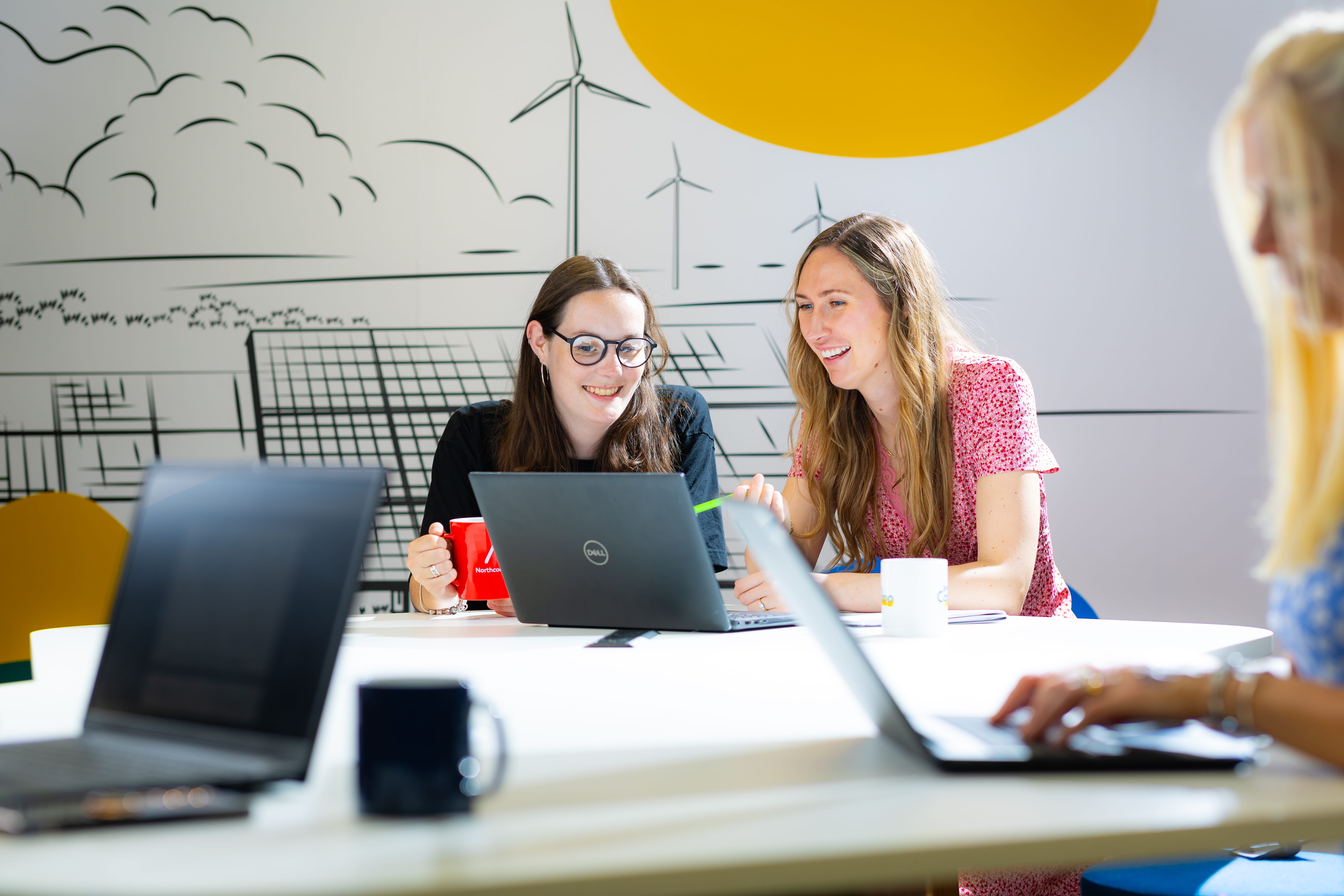 ALT - two ladies sitting at a desk working on laptops having a conversation with another lady typing on a laptop