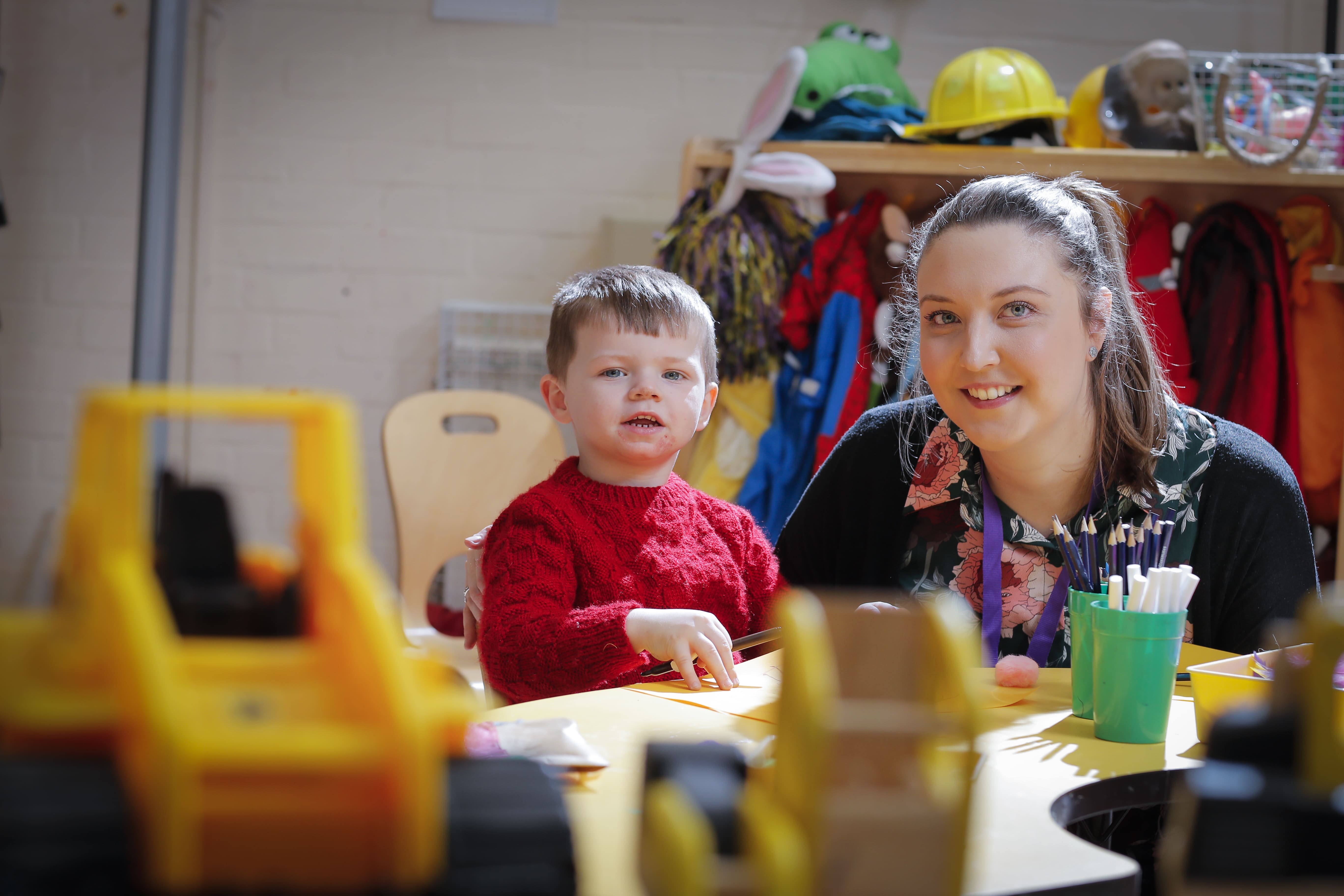 ALT - teacher and child sitting at a desk, both are smiling. Child has a pencil in his hand.