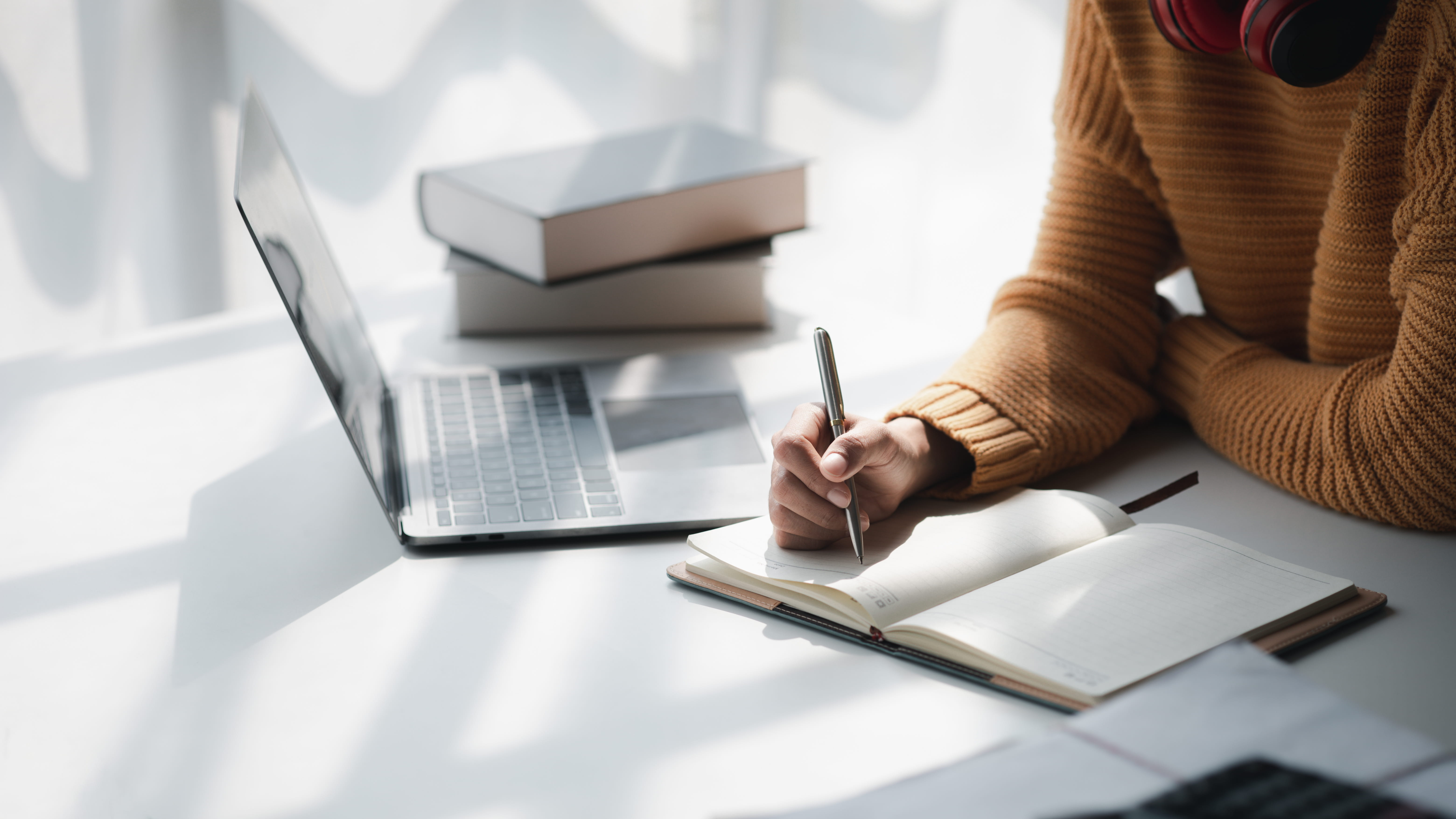 ALT - person writing with a pen in a notepad with a laptop beside them and some books