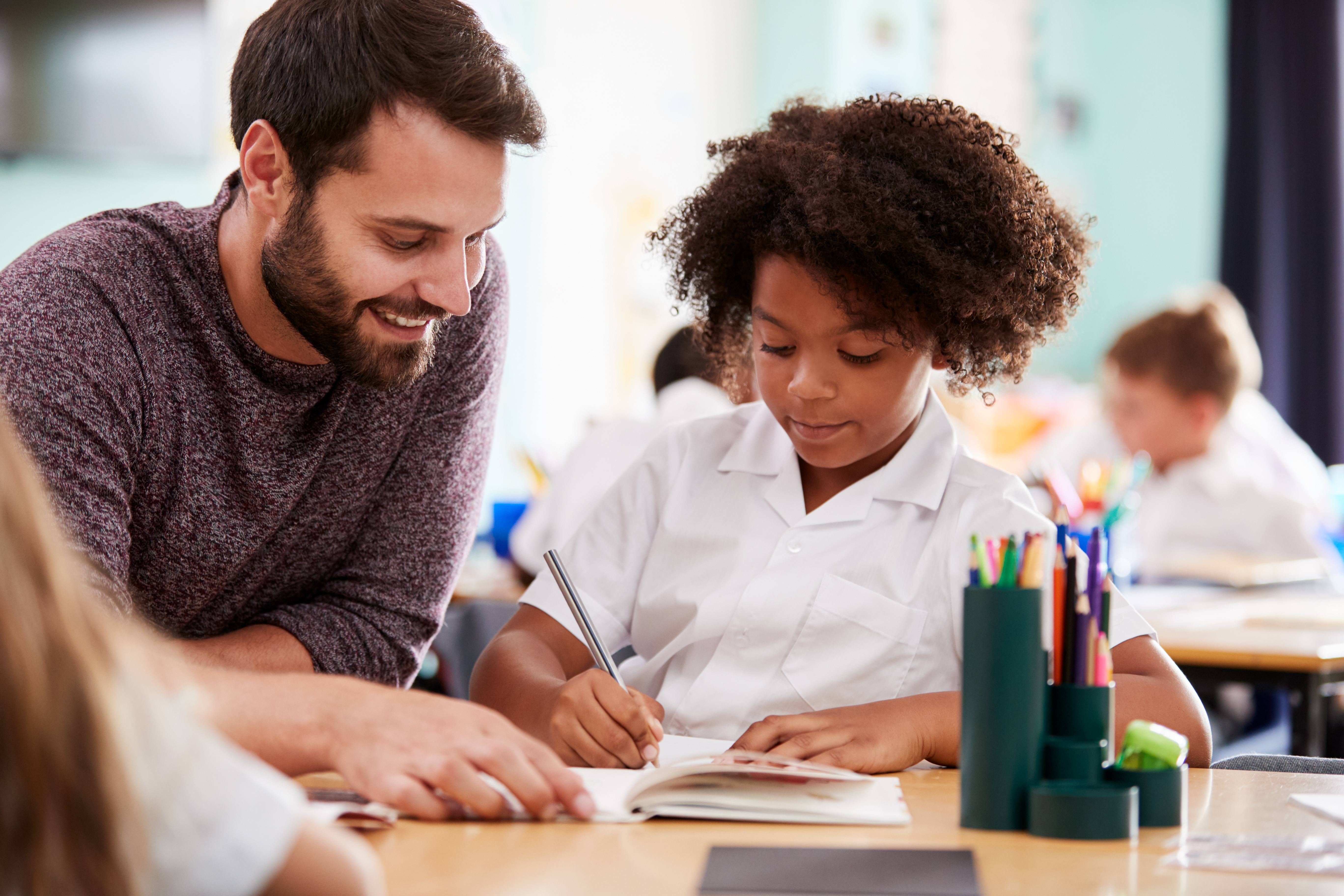 Alt - Teacher and child sitting at desk looking at an exercise book. Child has a pencil in his hand and is writing.