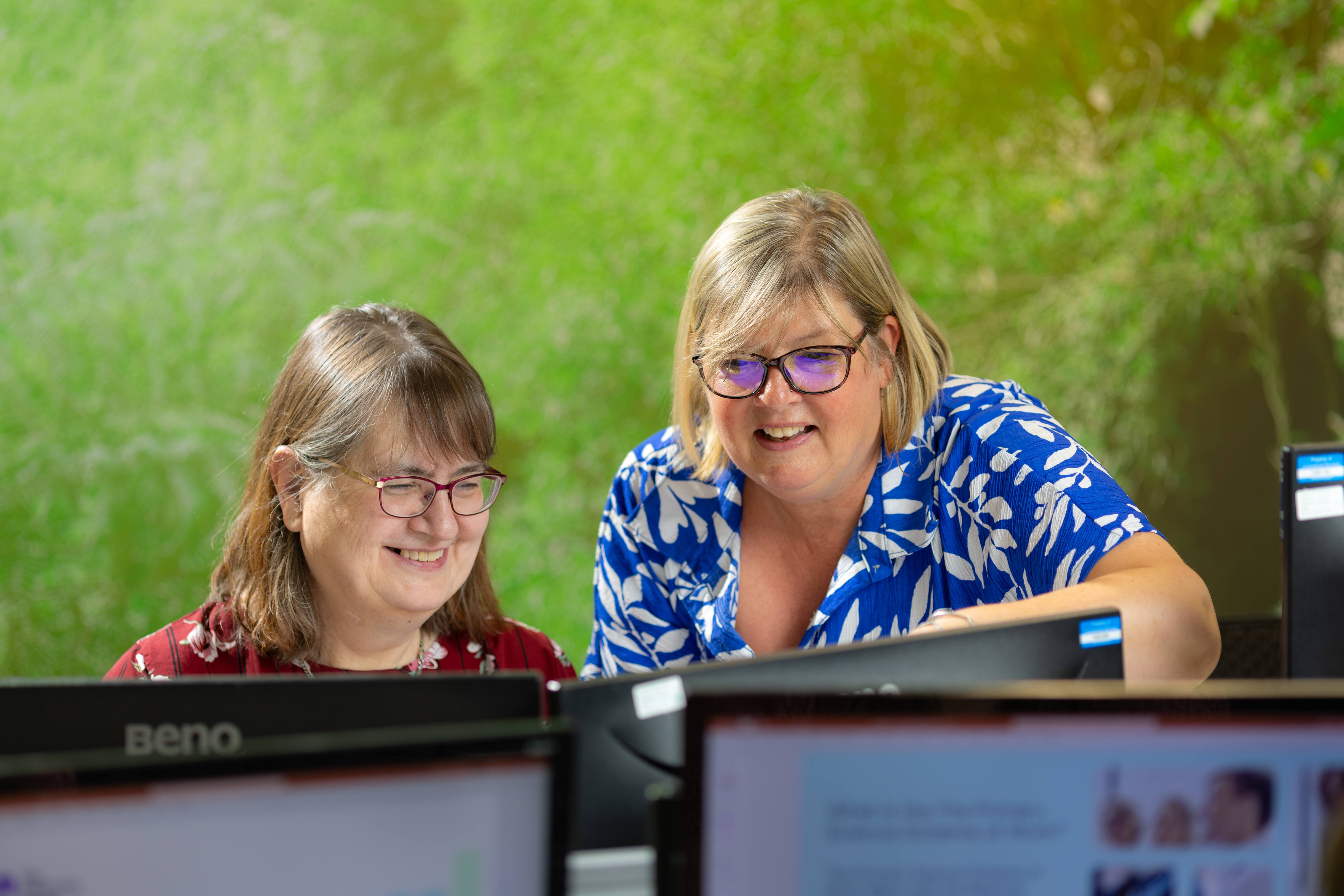  Two smiling women with glasses are sitting at a desk, engaged with computer screens. 