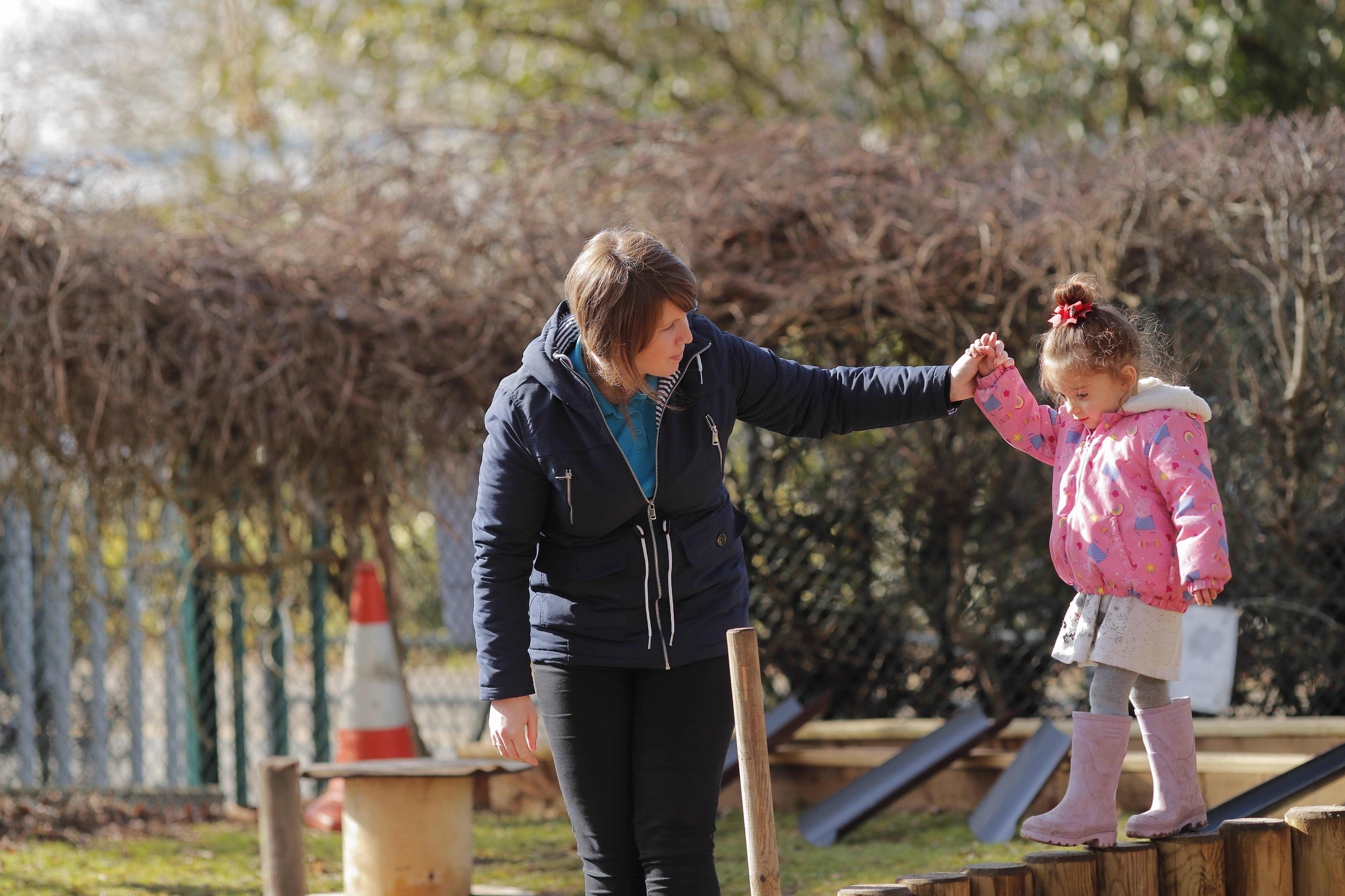 A woman helps a young girl balance on wooden beams outdoors. The girl wears a pink coat and boots, smiling, while the woman gently holds her hand.
