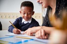 A young boy in a school uniform concentrates on writing with a pencil as a woman guides him, pointing at a notebook