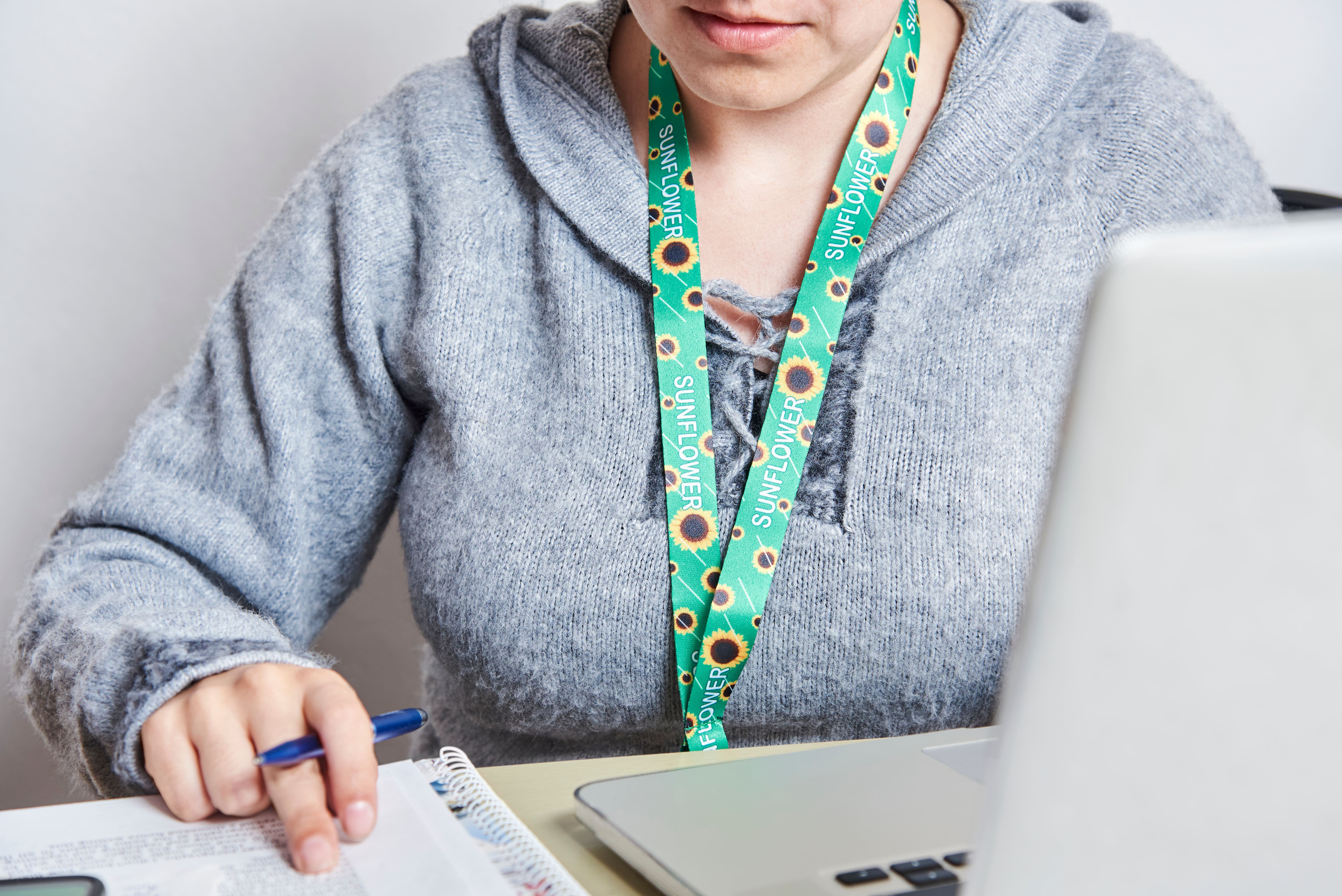 Person in grey sweater working at a desk with a laptop and papers. They wear a green sunflower lanyard.