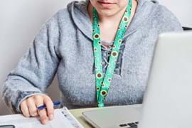 Person in grey sweater working at a desk with a laptop and papers. They wear a green sunflower lanyard.