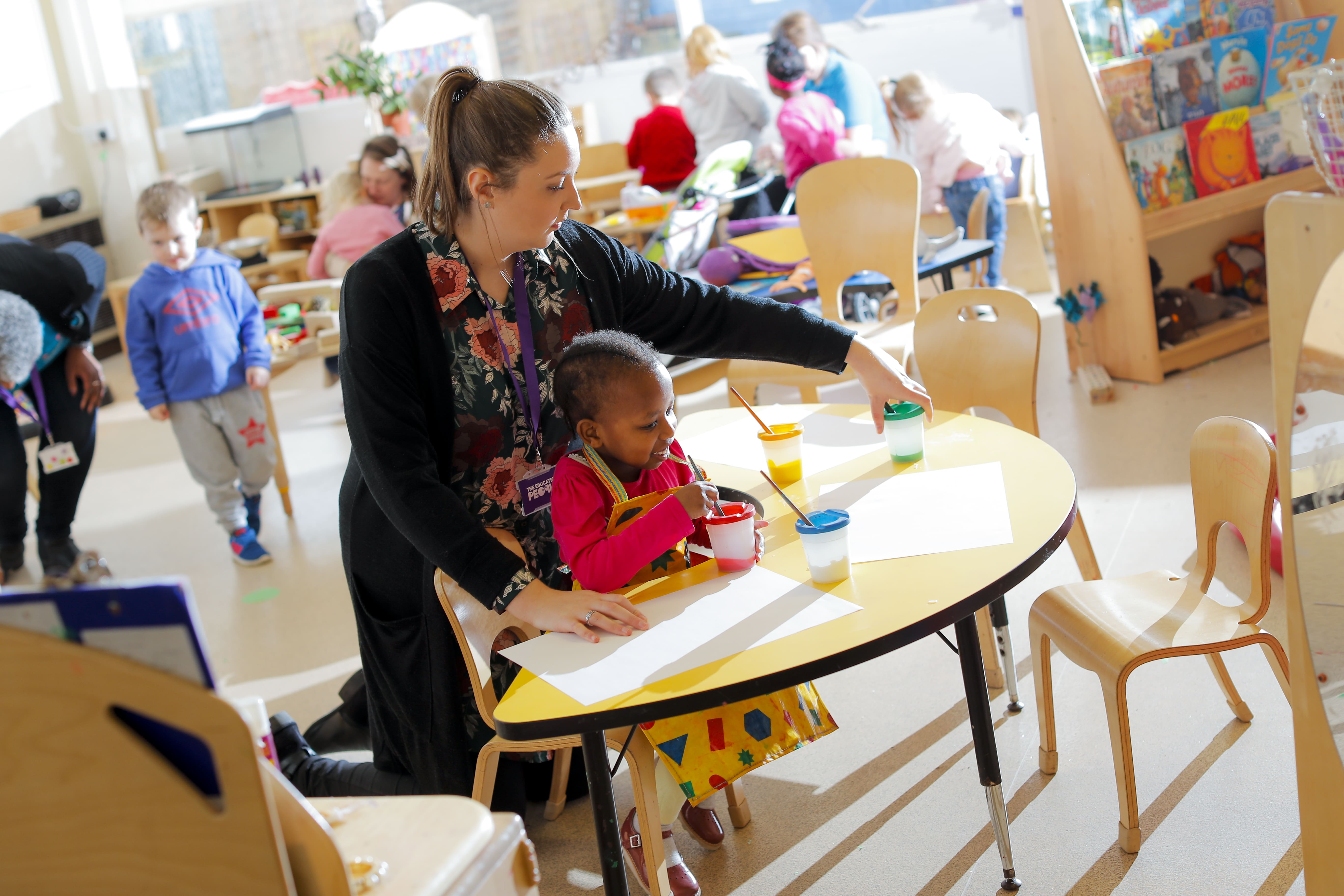 Nursery classroom with children playing, nursery teacher is helping a child to paint on paper on the table