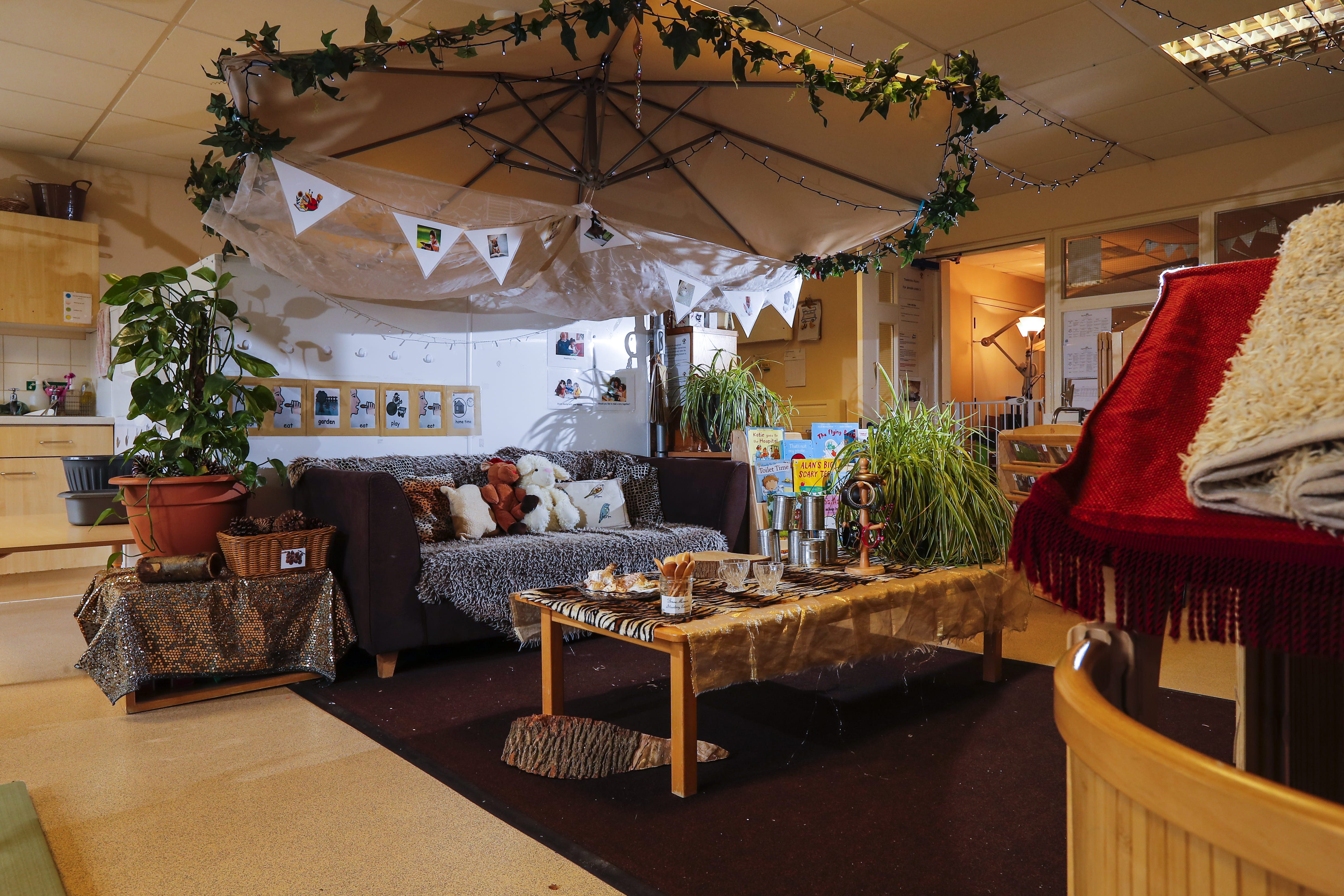 Nursery classroom with comfy seating cuddly toys, carpet, table and blankets with plants and flags hanging from the ceiling