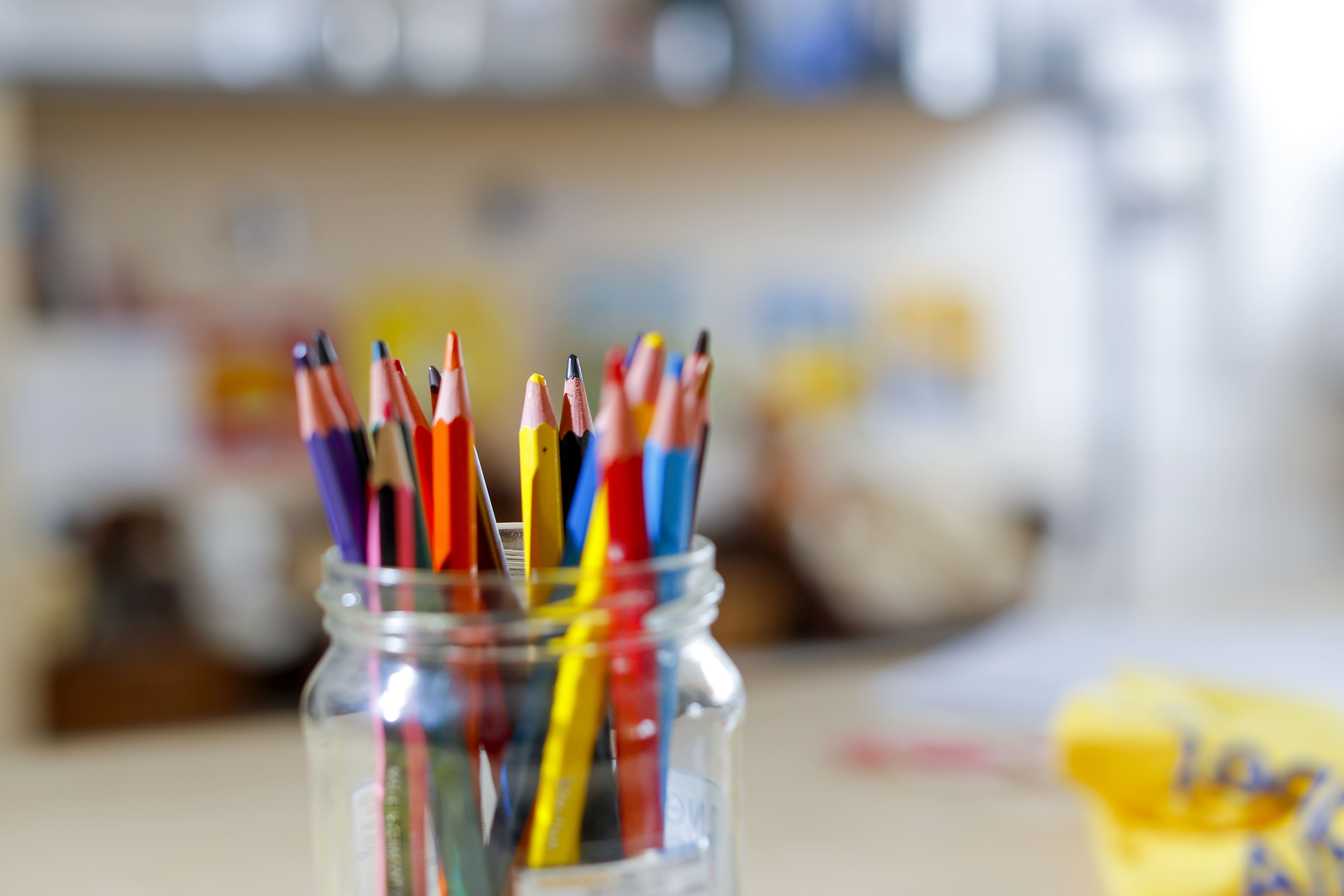 A mason jar filled with colourful pencils on a blurred desk, with a soft background of books and papers