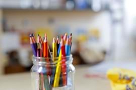 A mason jar filled with colourful pencils on a blurred desk, with a soft background of books and papers