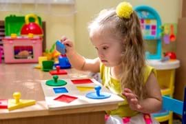 A young girl with a yellow flower in her hair is focused on fitting colourful geometric shapes into a wooden puzzle at a sunny playroom table.