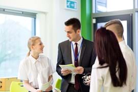 A teacher in a suit and lanyard talks to a group of students in school uniforms, holding notebooks. 