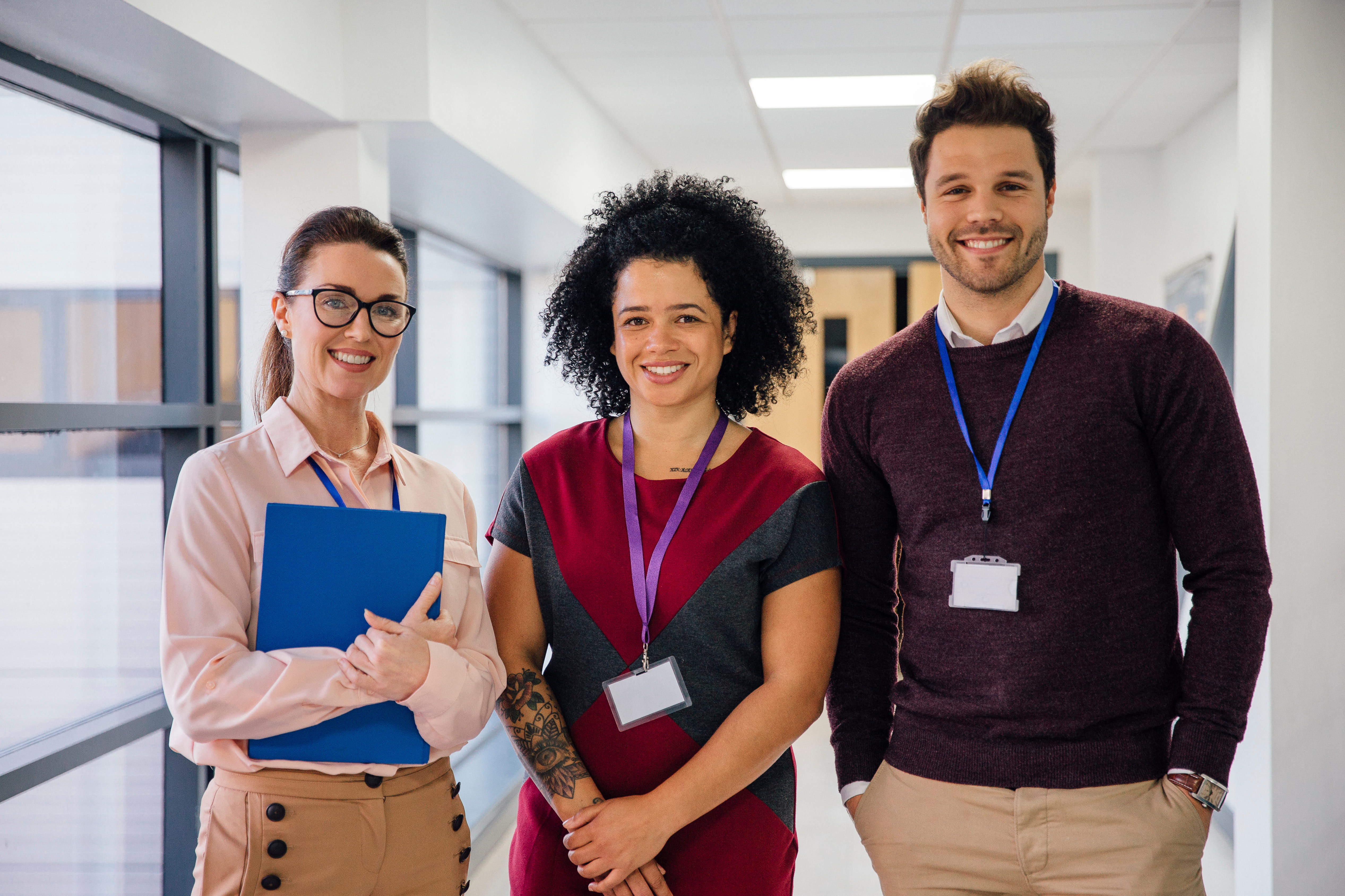Three colleagues smiling in a bright office hallway. One holds a blue folder, all wear ID badges.