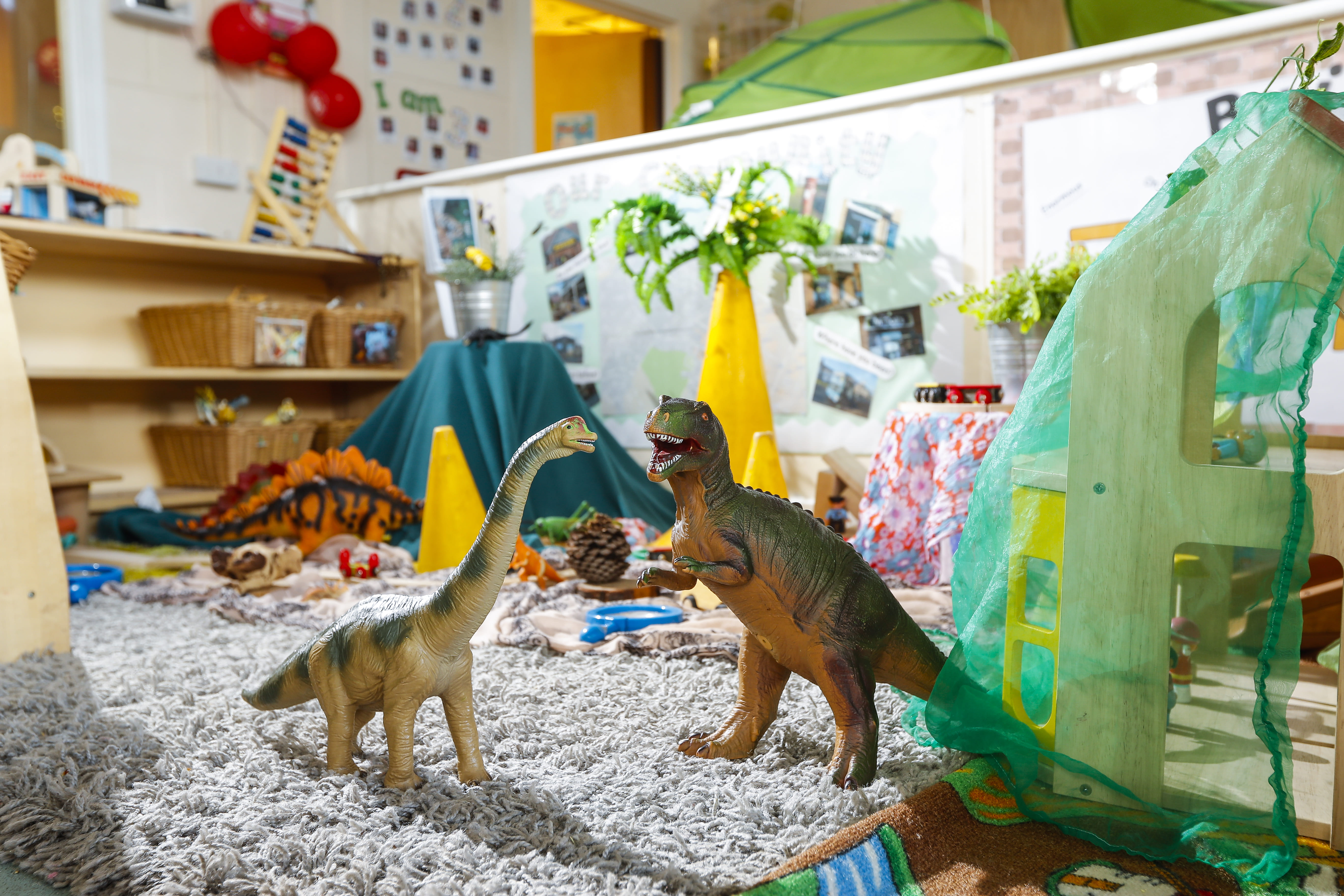 Toy dinosaurs, including a brachiosaurus and T-rex, stand on a playroom carpet.