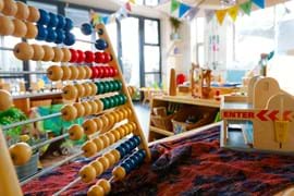 A nursery room scene with an abacus on top of a table covered in a blue and red cloth with a variety of toys in the distance