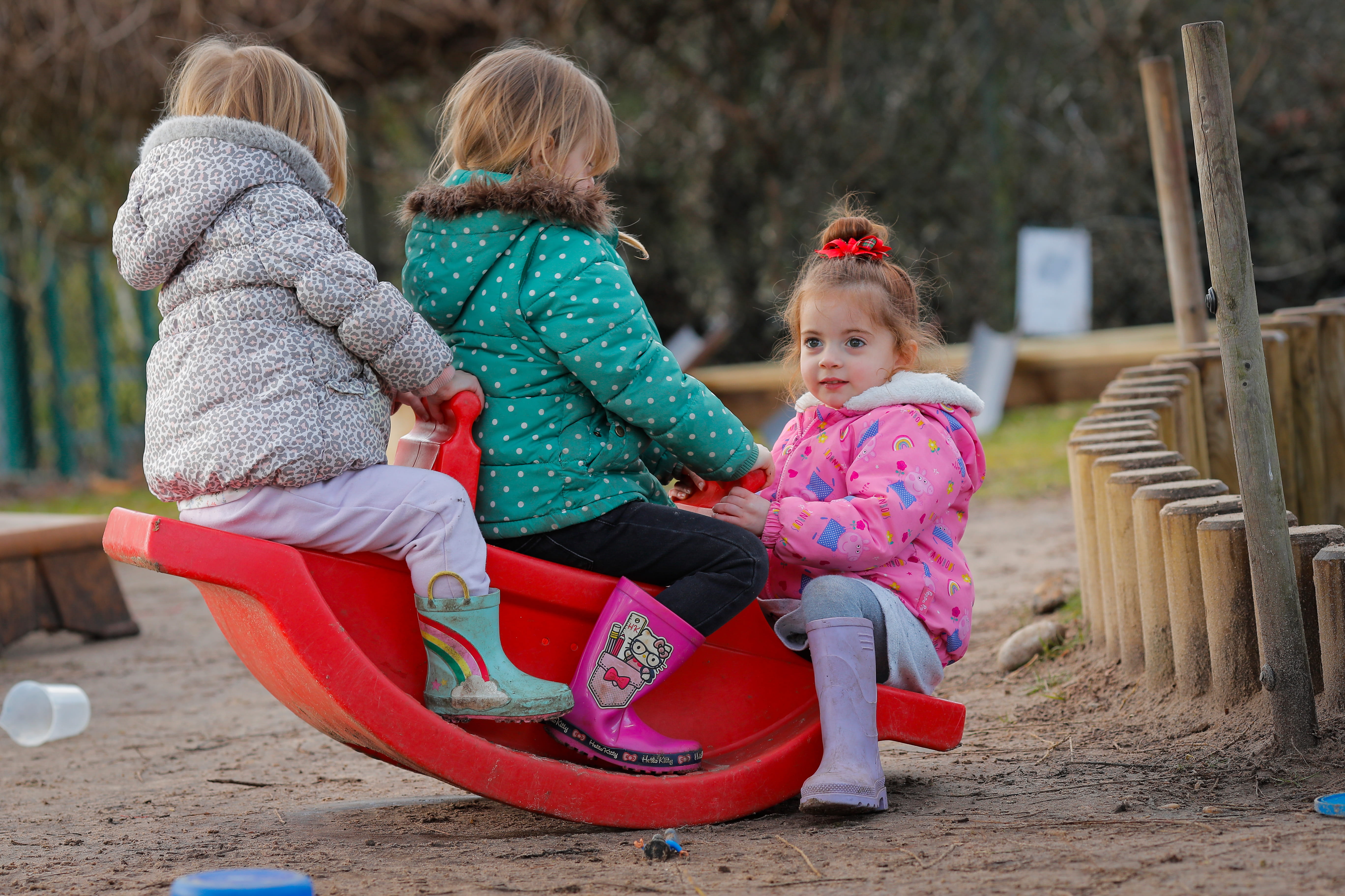 Three young children in colourful jackets and boots play happily on a red seesaw in a sandy park area, surrounded by wooden borders.
