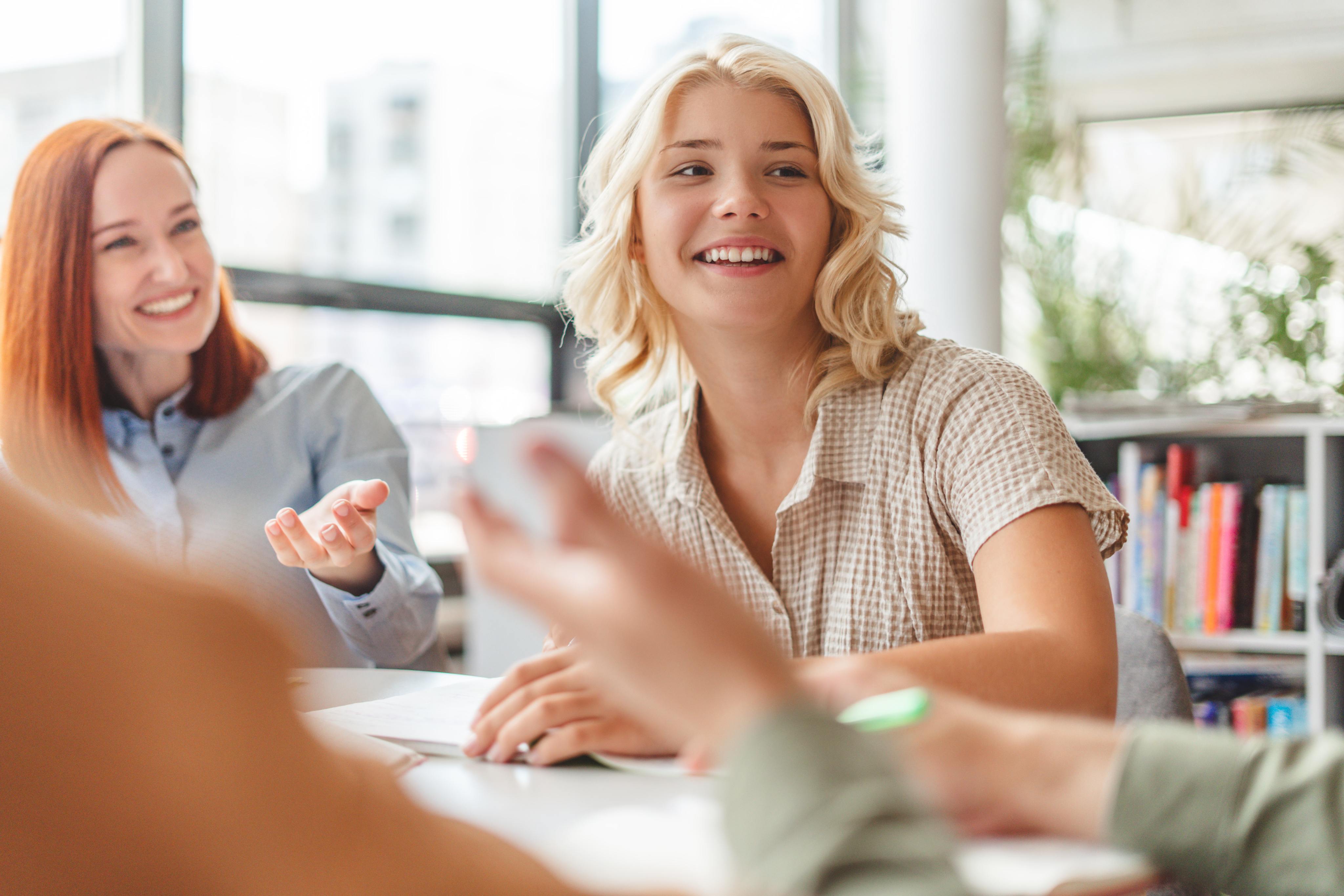 Two women smiling and conversing in a bright office setting. One has red hair and the other blonde.