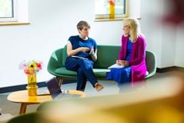 Two people sit on a green sofa in a well-lit room, engaged in deep conversation. A laptop and colourful flowers are on nearby tables