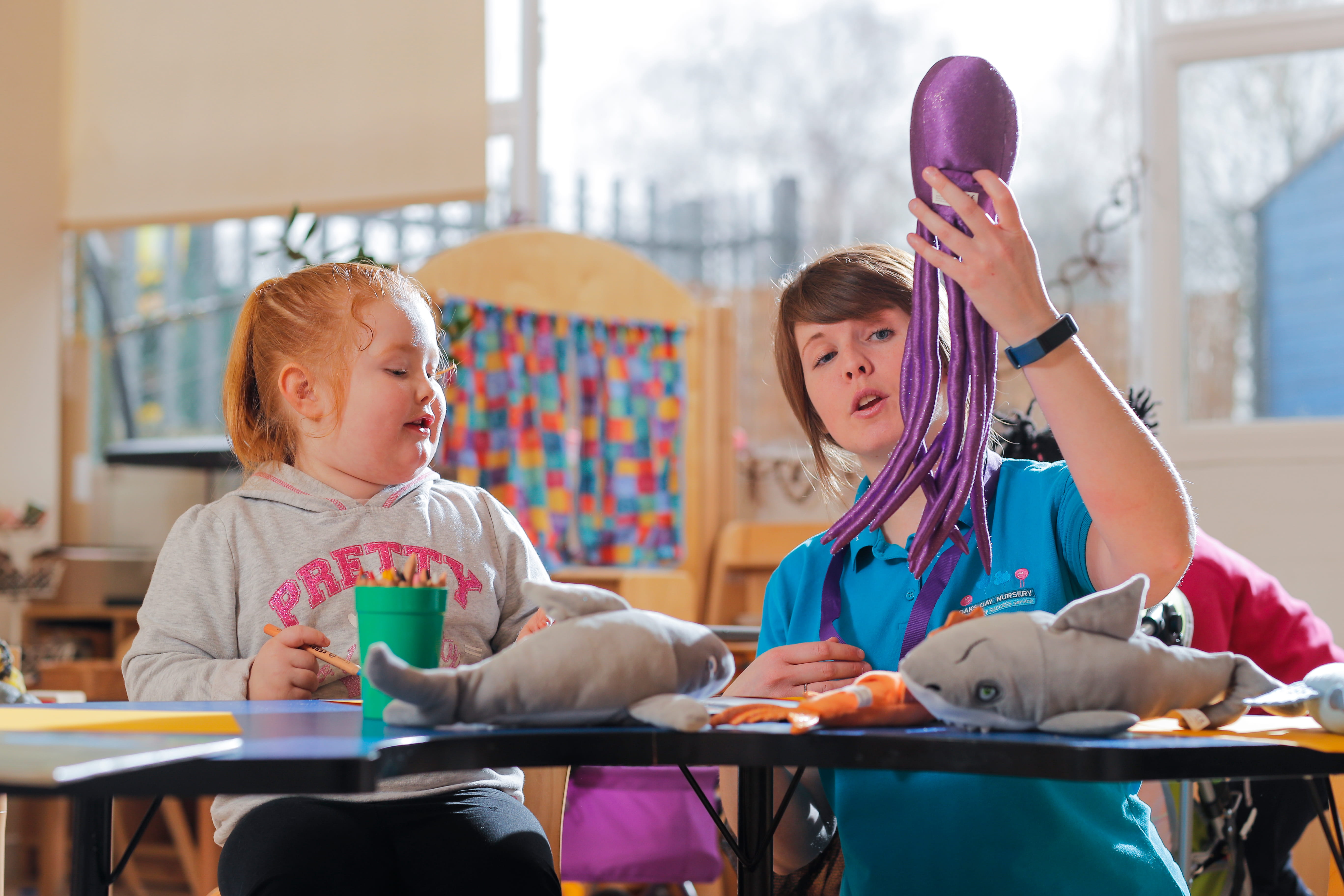 A nursery teacher helps a child with crafts at a table, surrounded by toy sharks. The teacher is holding up a toy octopus and talking about it.