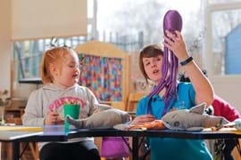 A nursery teacher helps a child with crafts at a table, surrounded by toy sharks. The teacher is holding up a toy octopus and talking about it.