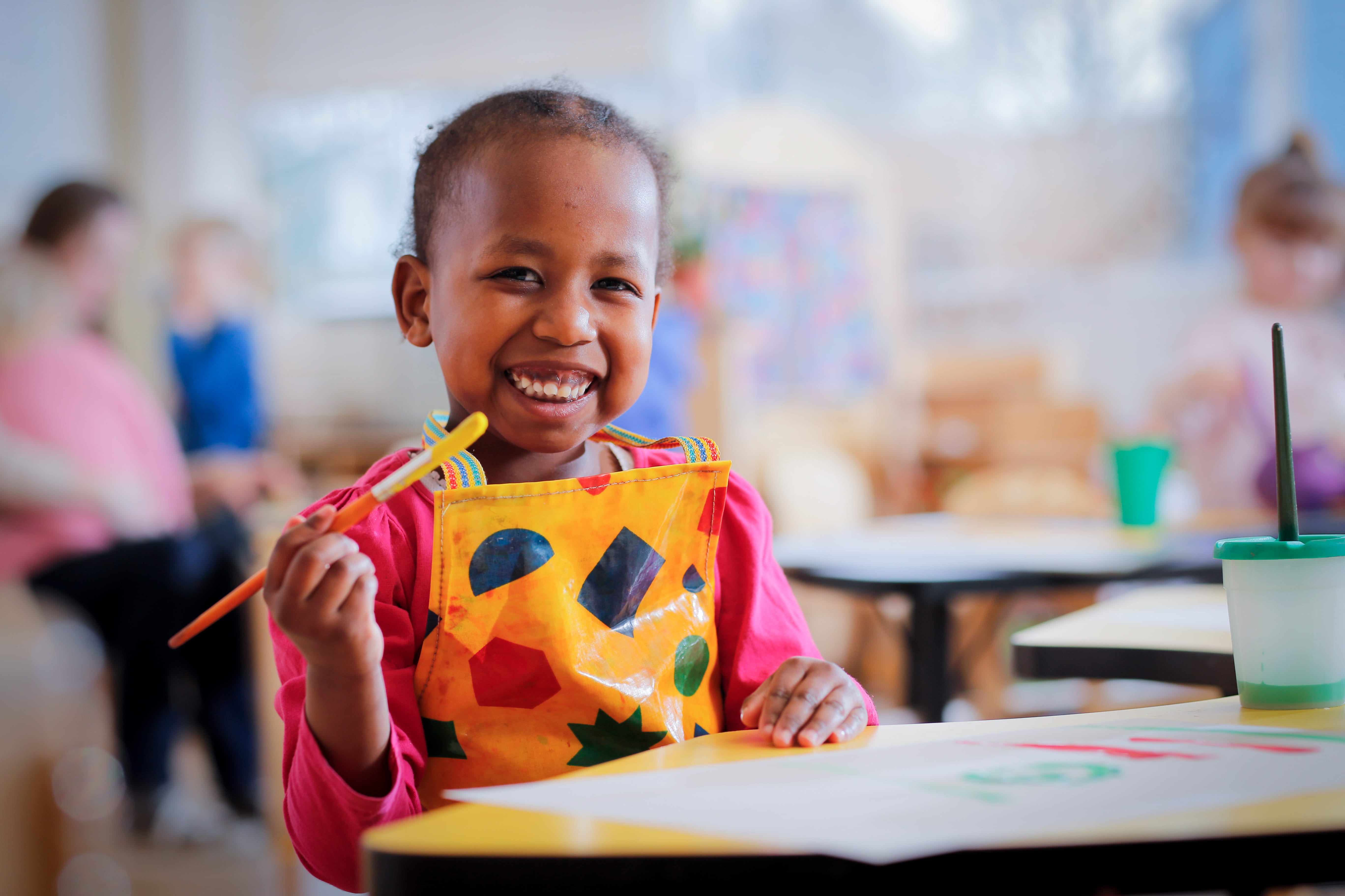 Smiling child in a classroom, wearing a colourful apron holds a paintbrush.